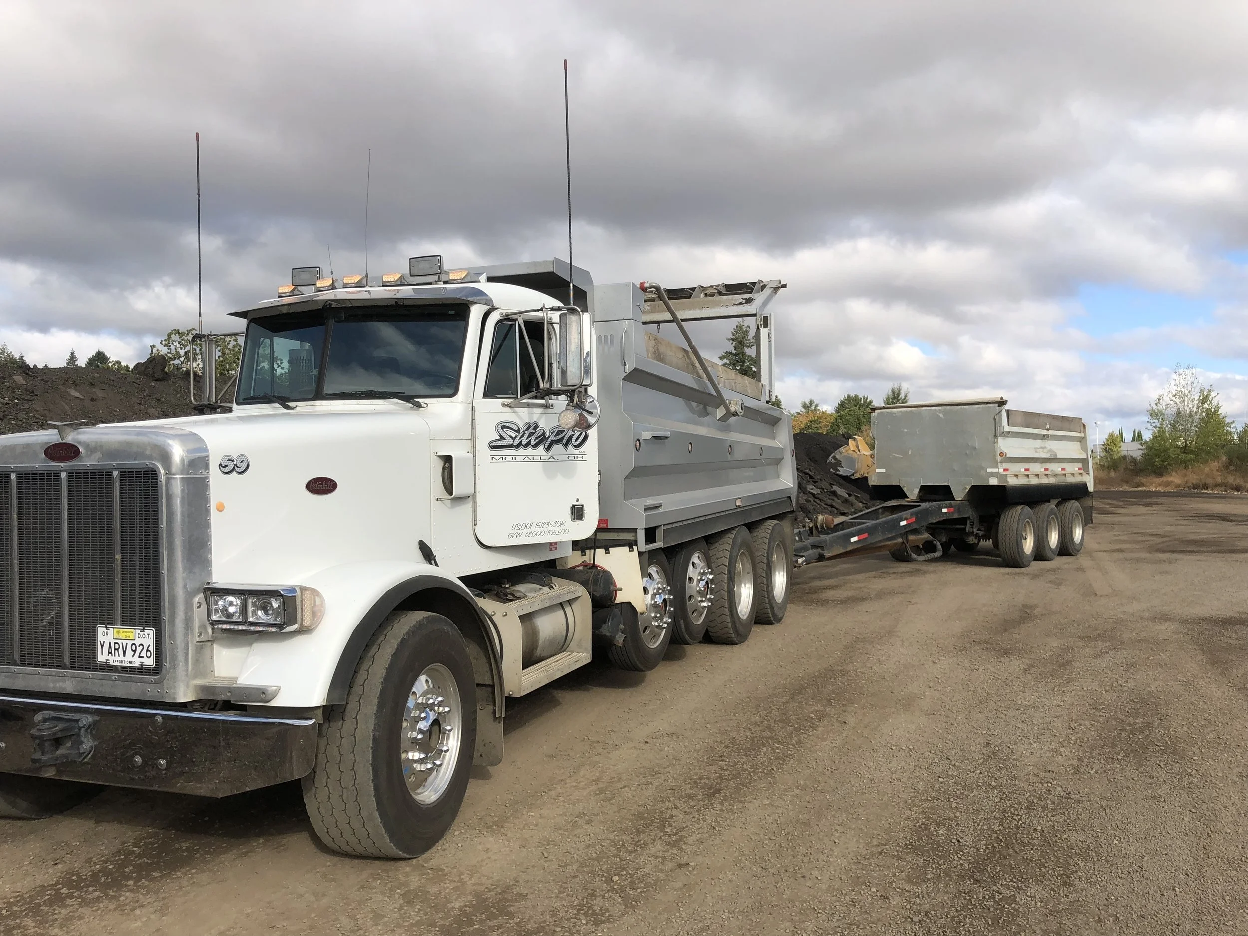 A white Peterbilt semi-truck with a dump trailer attached, parked on a dirt lot under a cloudy sky.
