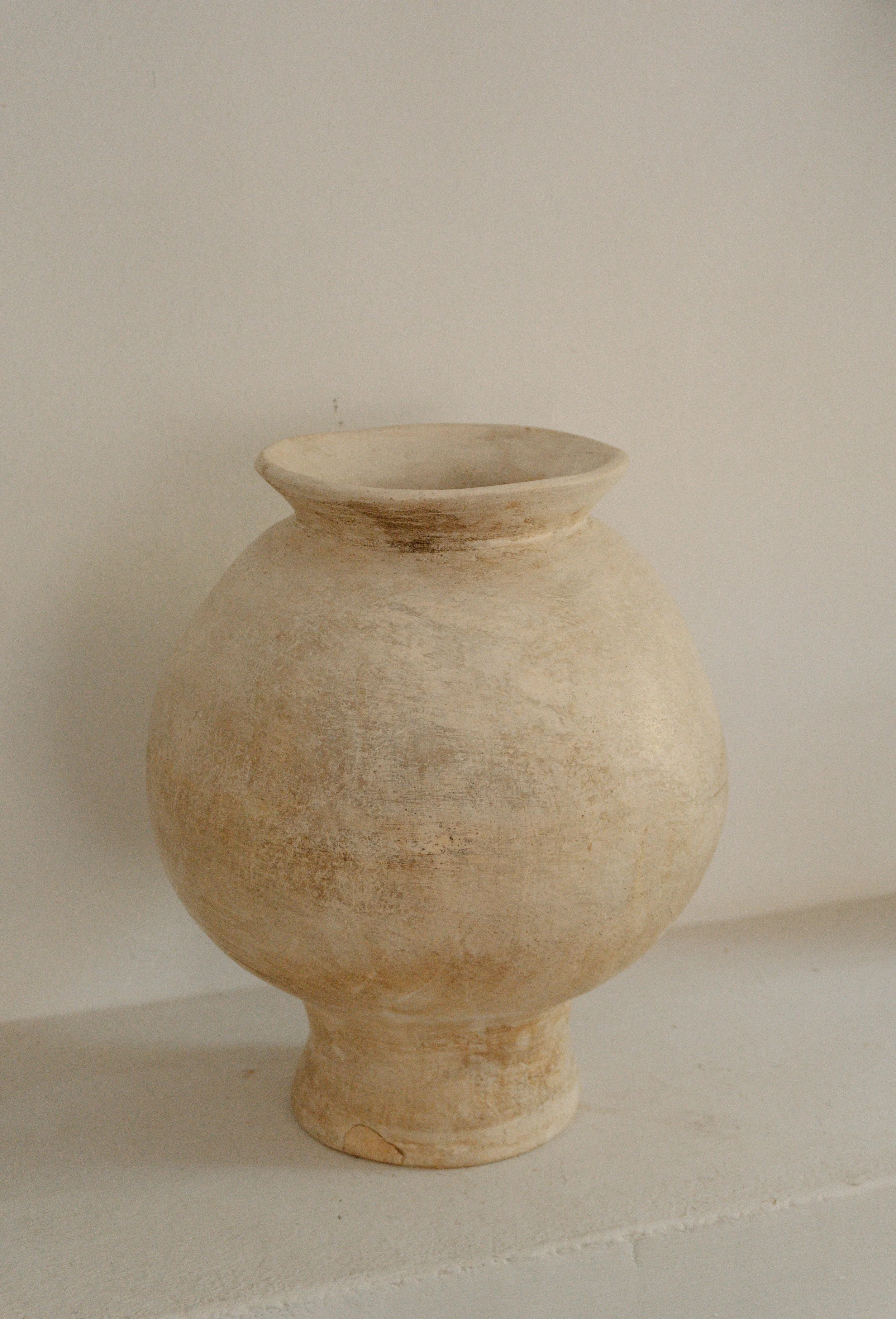 Ancient beige clay vase with a narrow base and wide round body, set against a plain off-white background.
