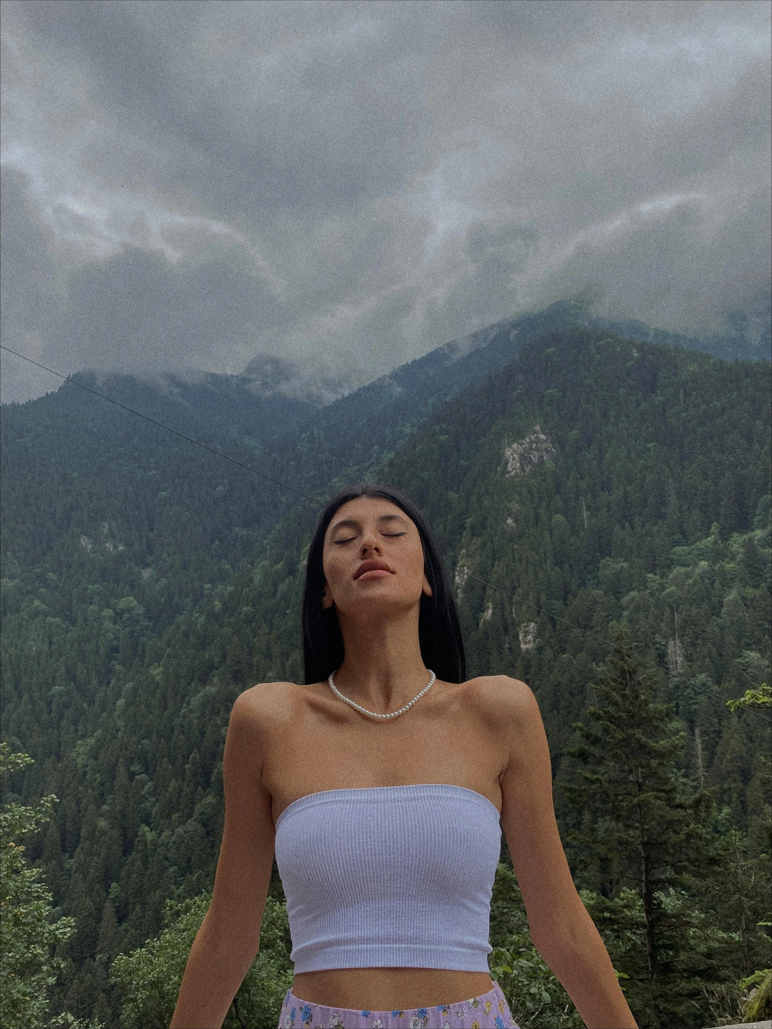 A woman with long black hair and closed eyes stands outdoors with a mountain and cloudy sky in the background. She is wearing a strapless white top and a pearl necklace.