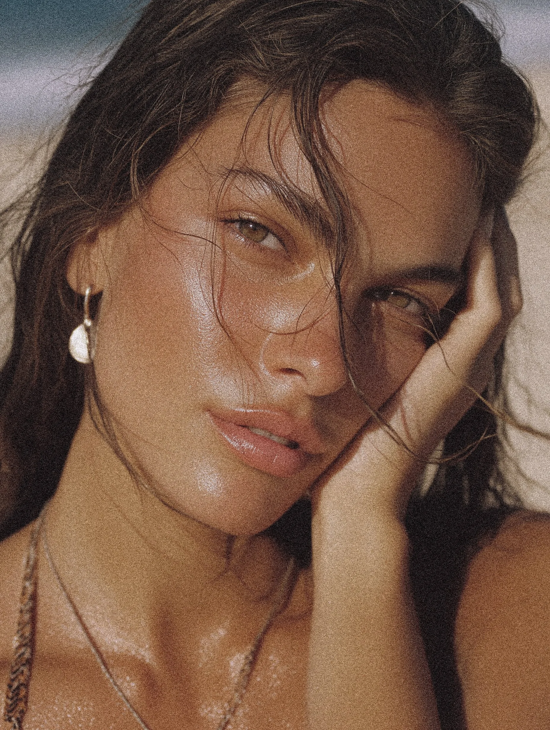 A close-up portrait of a woman with wet hair, minimal makeup, and wearing earrings and necklaces, resting her face on her hand.