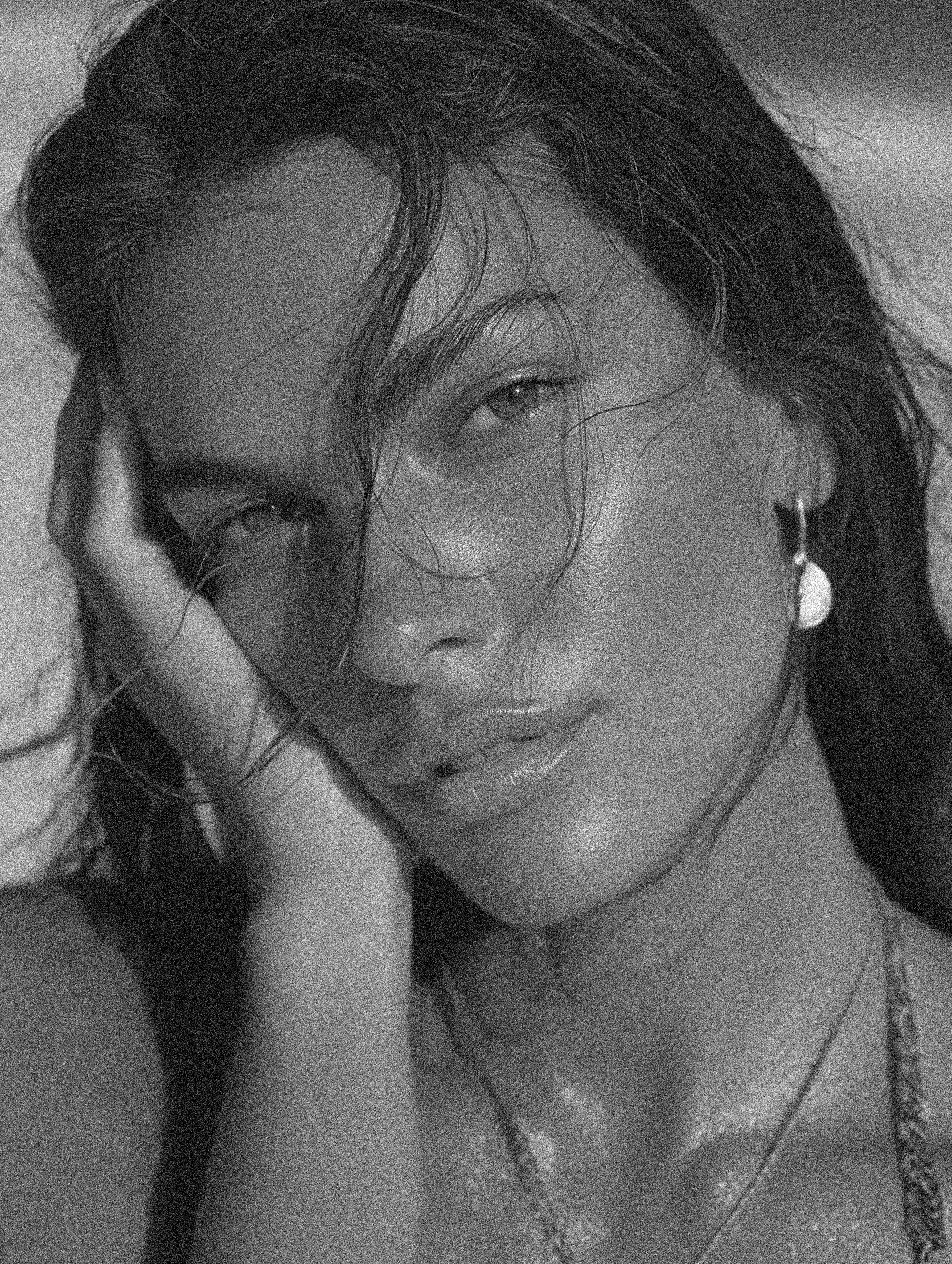 A black-and-white close-up photo of a woman with wavy hair, smoky eye makeup, and glossy lips, resting her face on her hand with a relaxed expression.