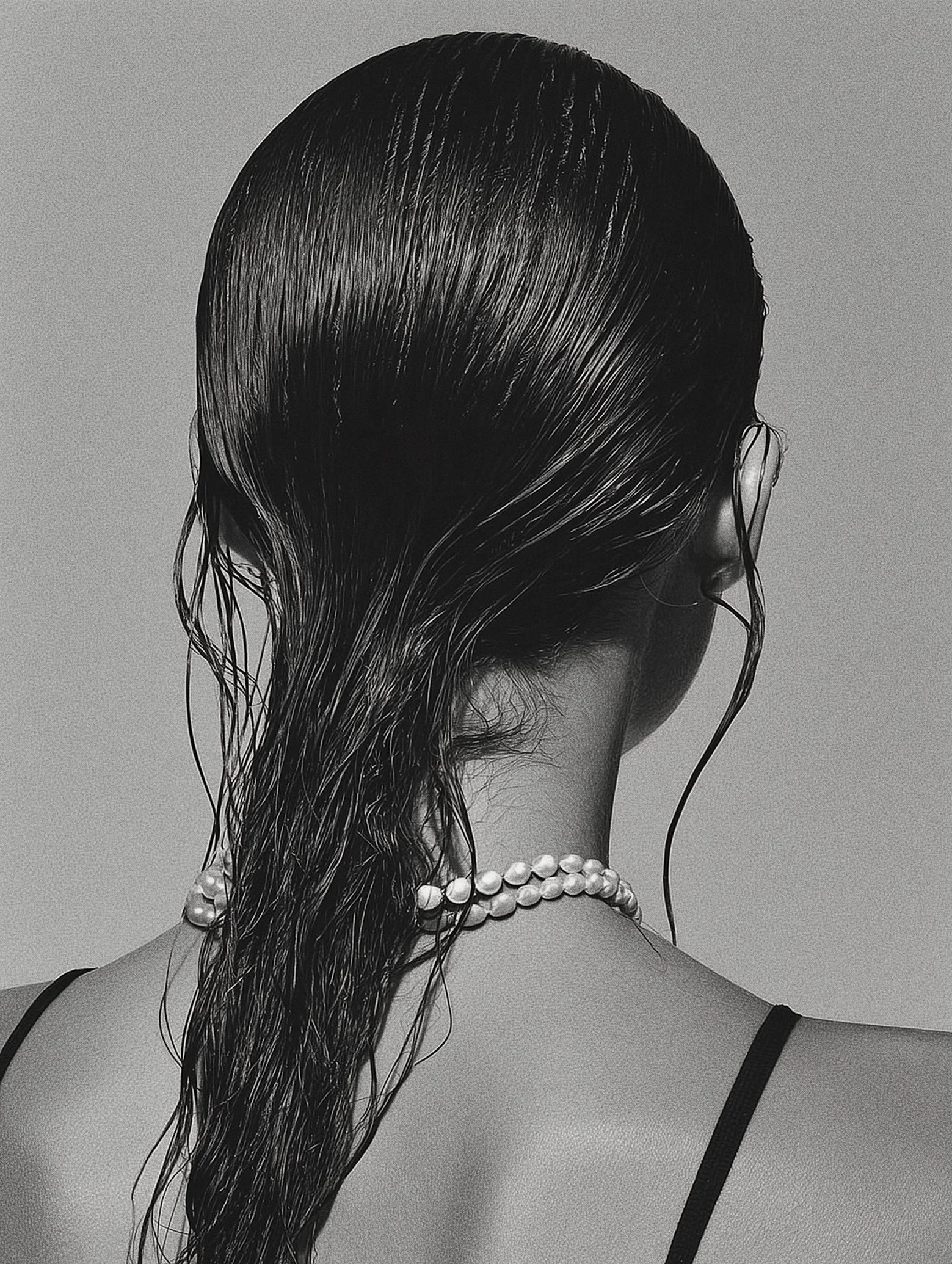 A black and white photo of a woman with wet, slicked-back hair, a pearl necklace, and a spaghetti strap top, viewed from behind.