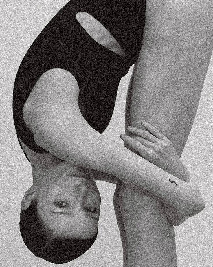 A black and white photograph of a woman holding her leg with her hand, her head upside-down and looking at the camera. She has short hair and a small tattoo on her wrist.