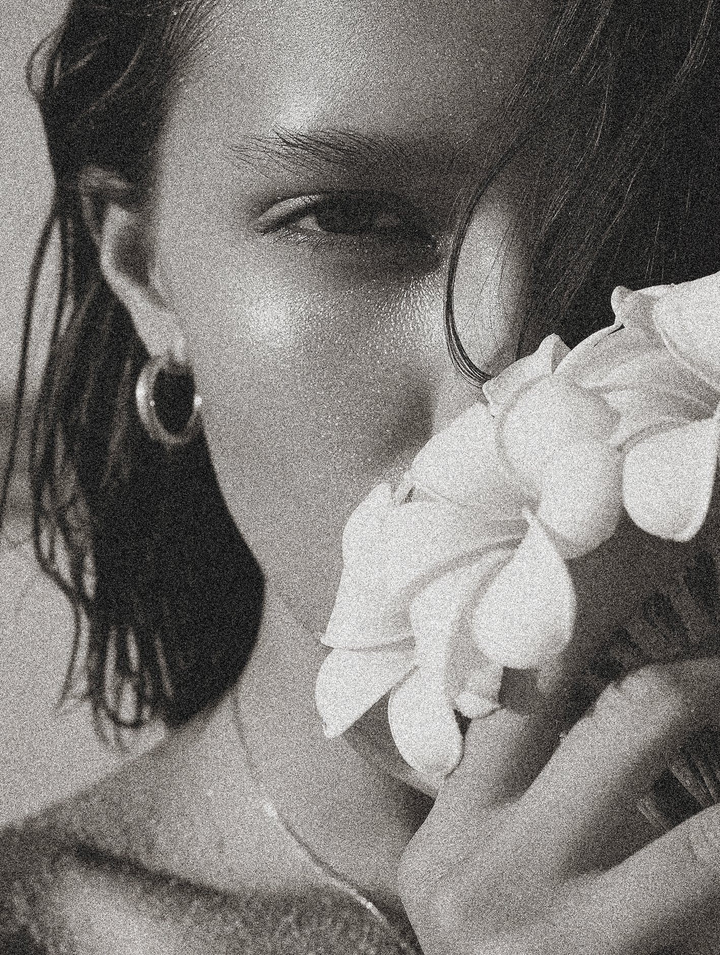 A close-up black and white photograph of a woman holding a flower near her face, with visible earrings and slightly wavy hair.