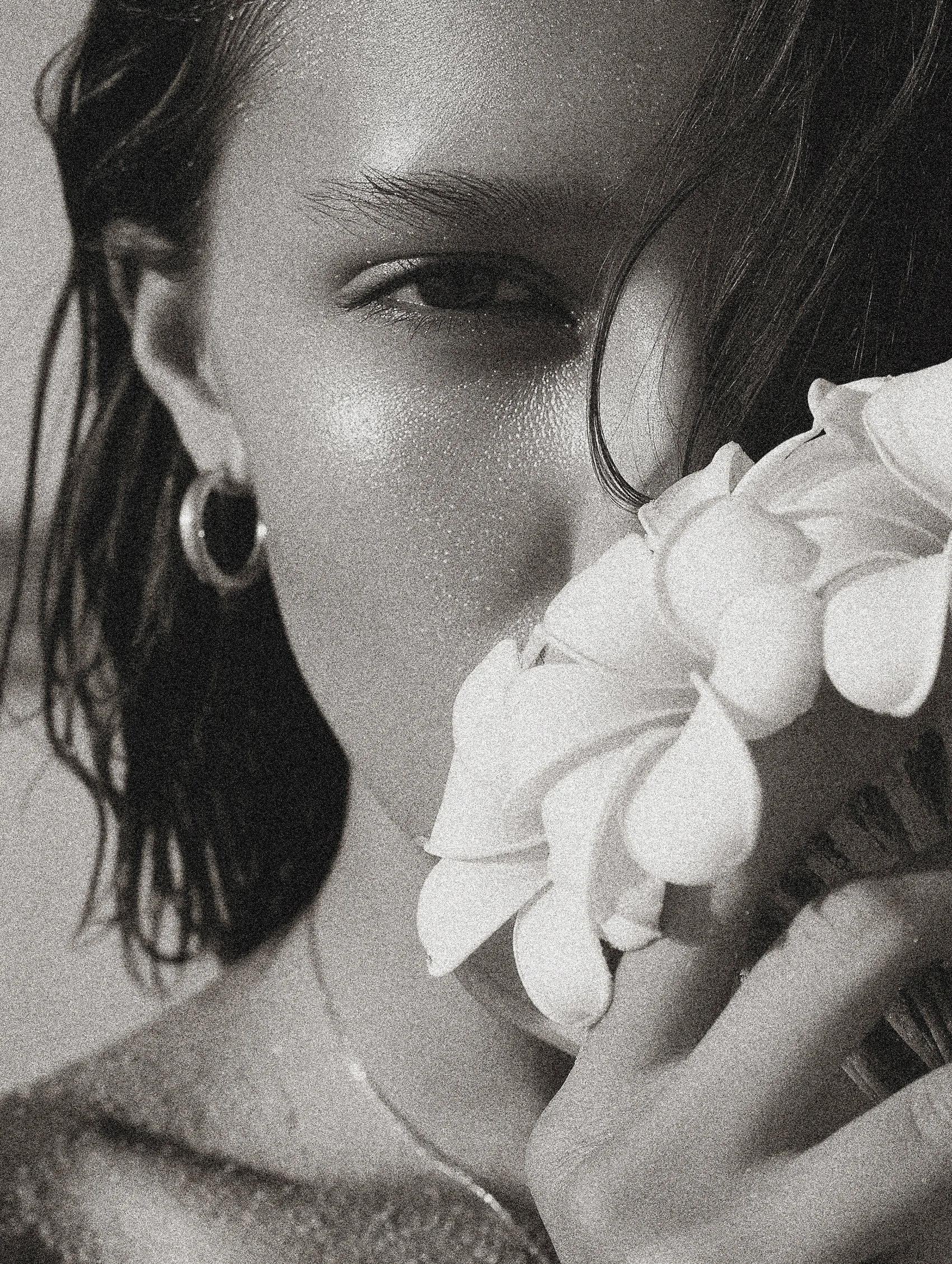 Close-up black and white photo of a woman holding a white flower near her face, partially covering her mouth, with textured skin and wet hair, wearing hoop earrings.