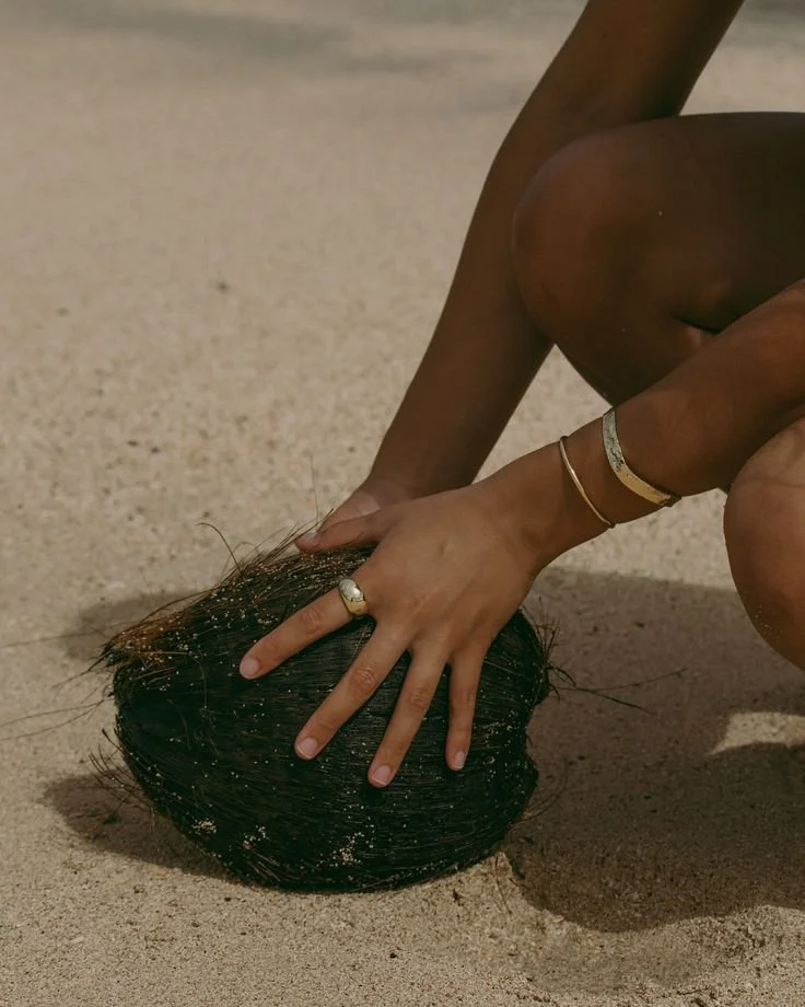 A person crouching on the sand at the beach, touching a large, dark, shiny, round object with seaweed and sand on it, wearing rings and bracelets.