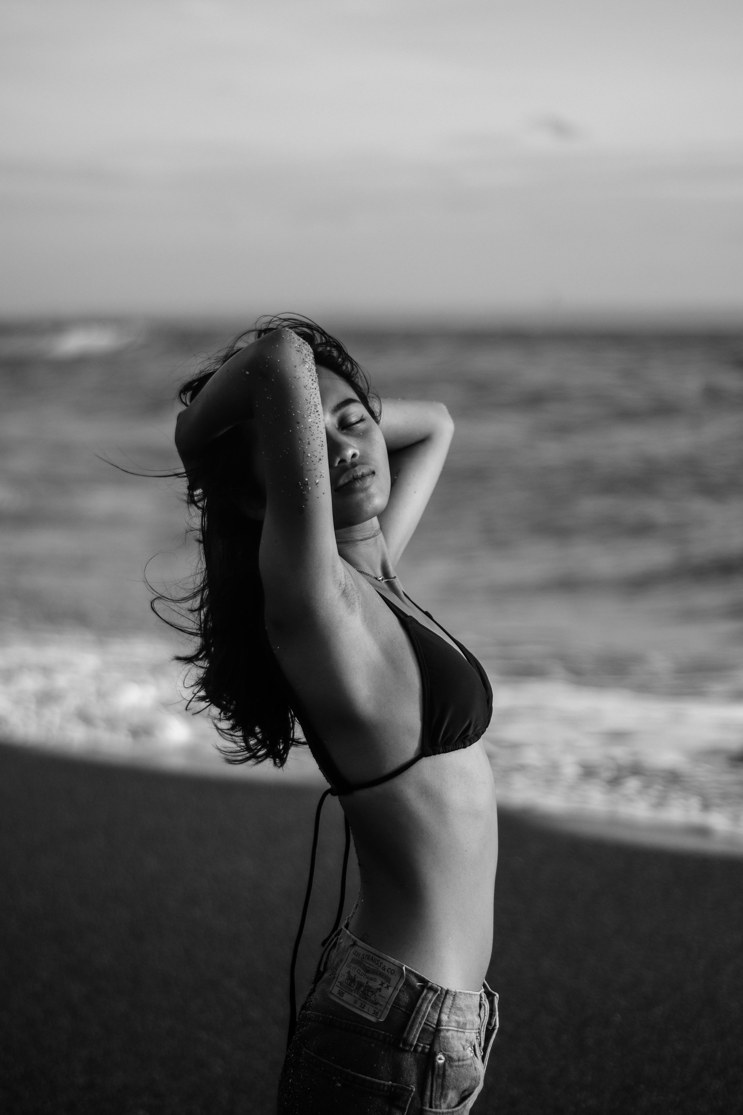 Black and white photo of a woman in a bikini top and shorts on the beach with her eyes closed and arms raised behind her head, looking relaxed.