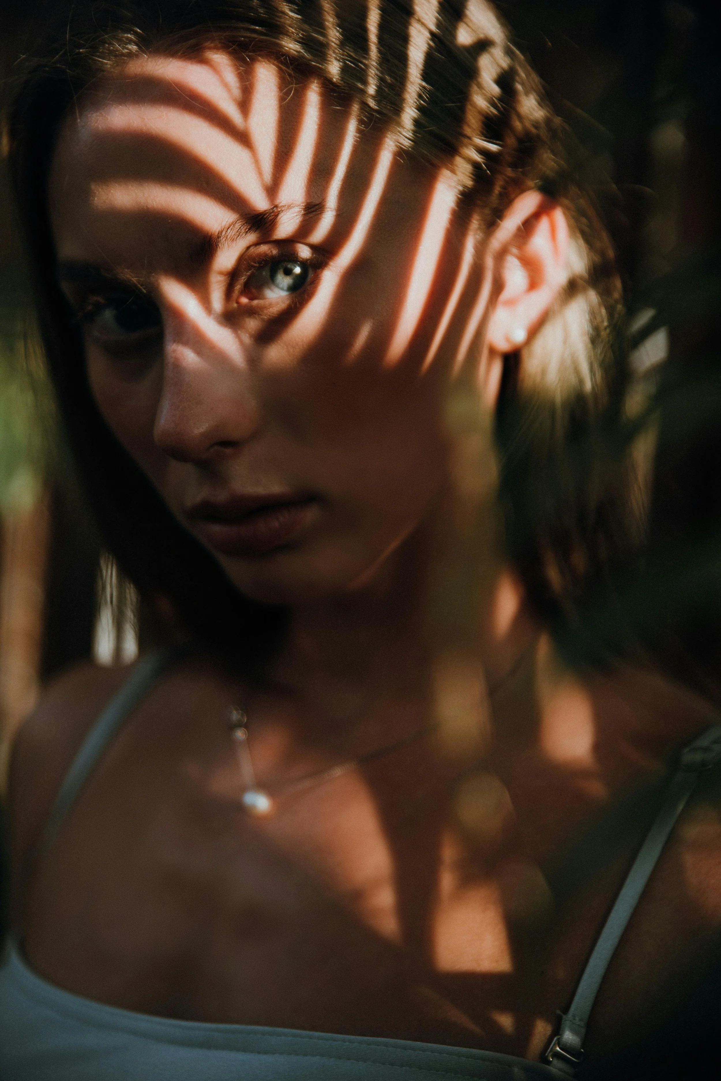A young woman outdoors with sunlight creating striped shadows on her face through leaves, wearing a light-colored top and a delicate necklace.
