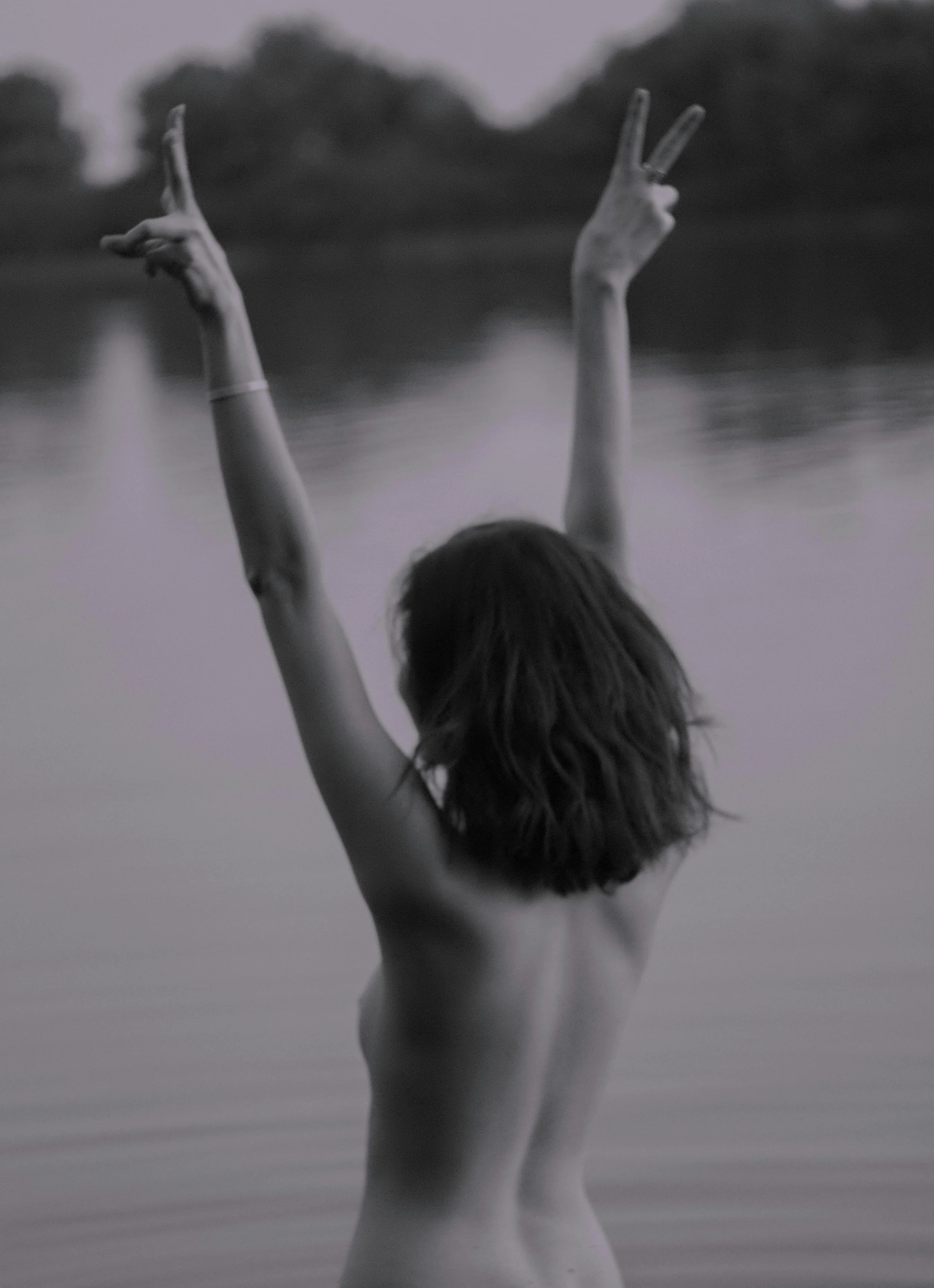 A woman with shoulder-length hair, seen from behind, is standing nude in a body of water with her arms raised, making peace signs with both hands, against a blurred natural background.