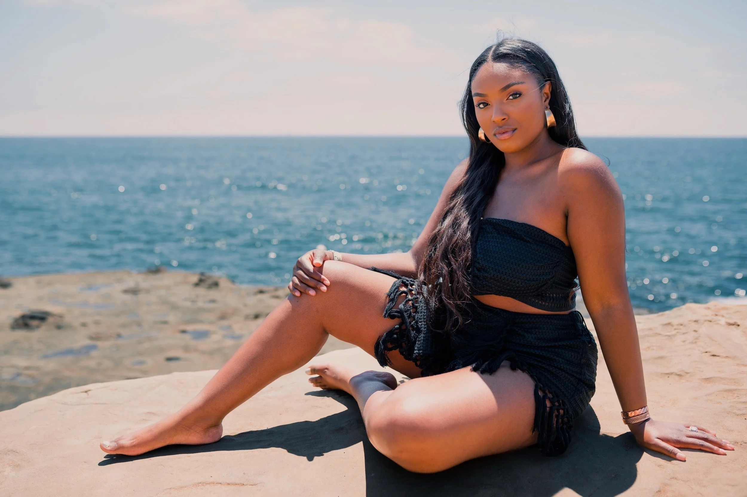 A woman sitting on a rock by the ocean, wearing a black strapless top and shorts, with long dark hair and earrings.