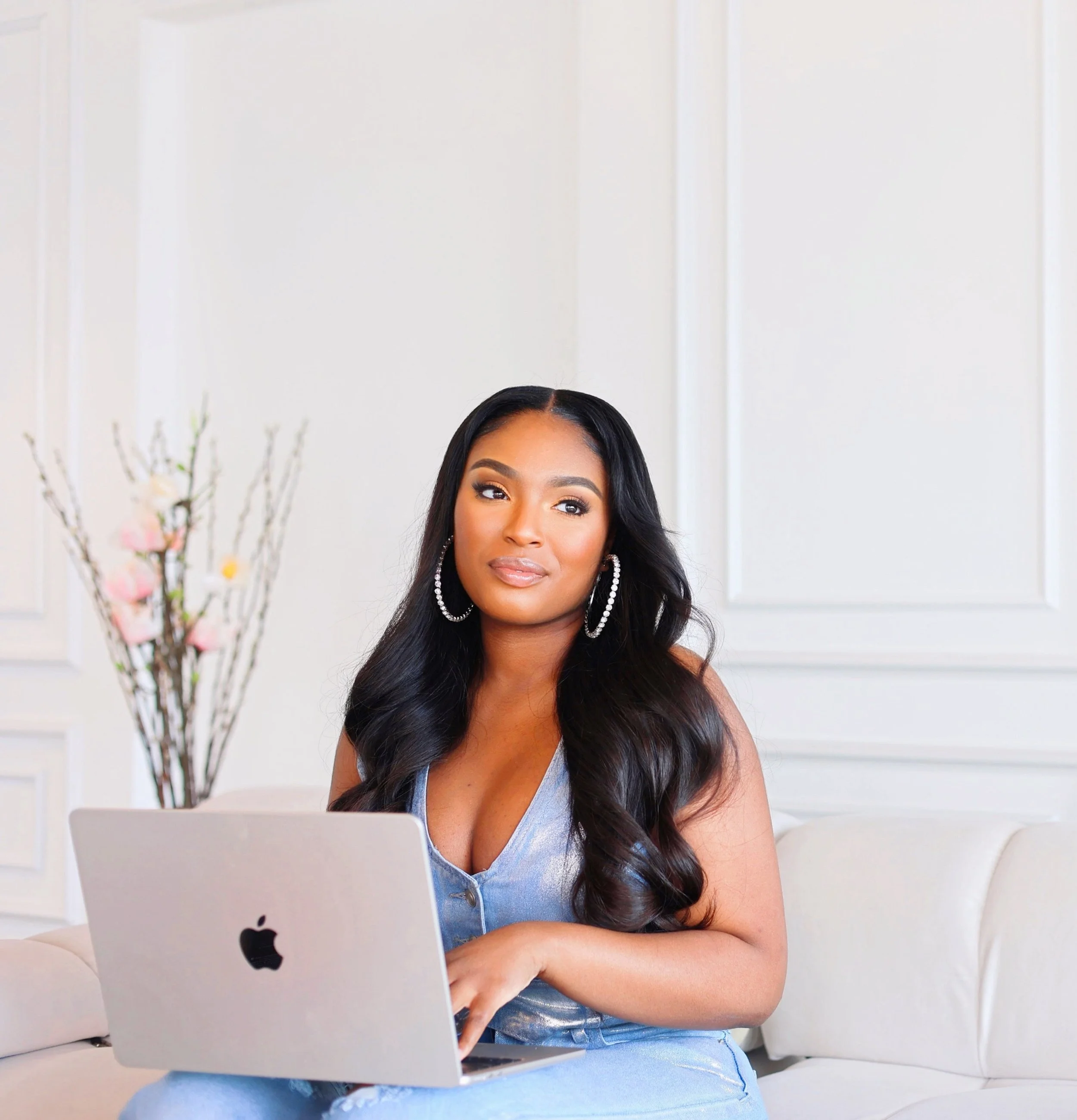 A woman with long black hair and large hoop earrings sitting on a cream-colored sofa, using a silver MacBook laptop, in a bright room with white walls and a decorative branch with pink flowers in the background.