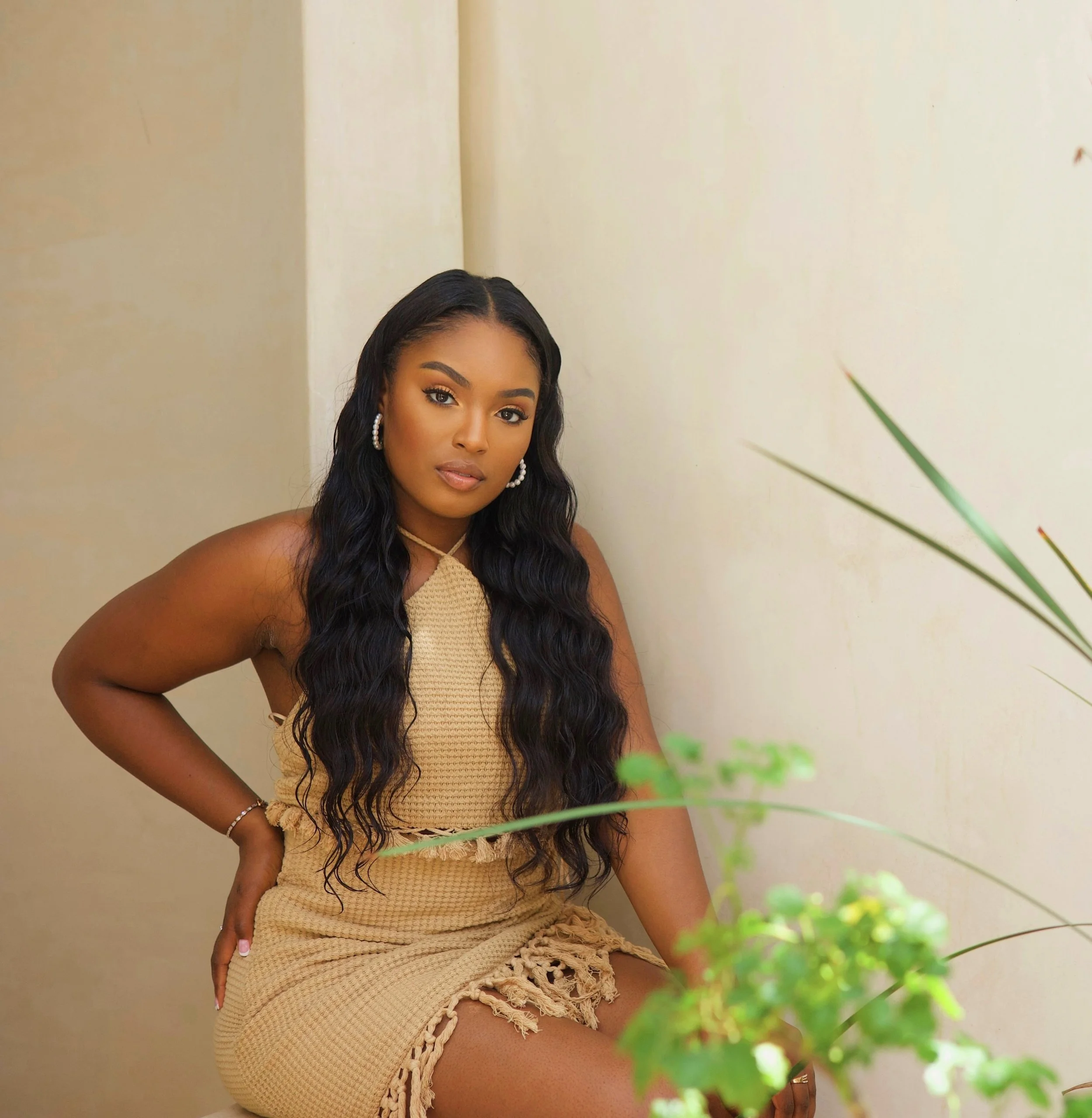 A young woman with long, wavy black hair, wearing a beige sleeveless dress and pearl earrings, sitting indoors near a wall with a plant nearby.
