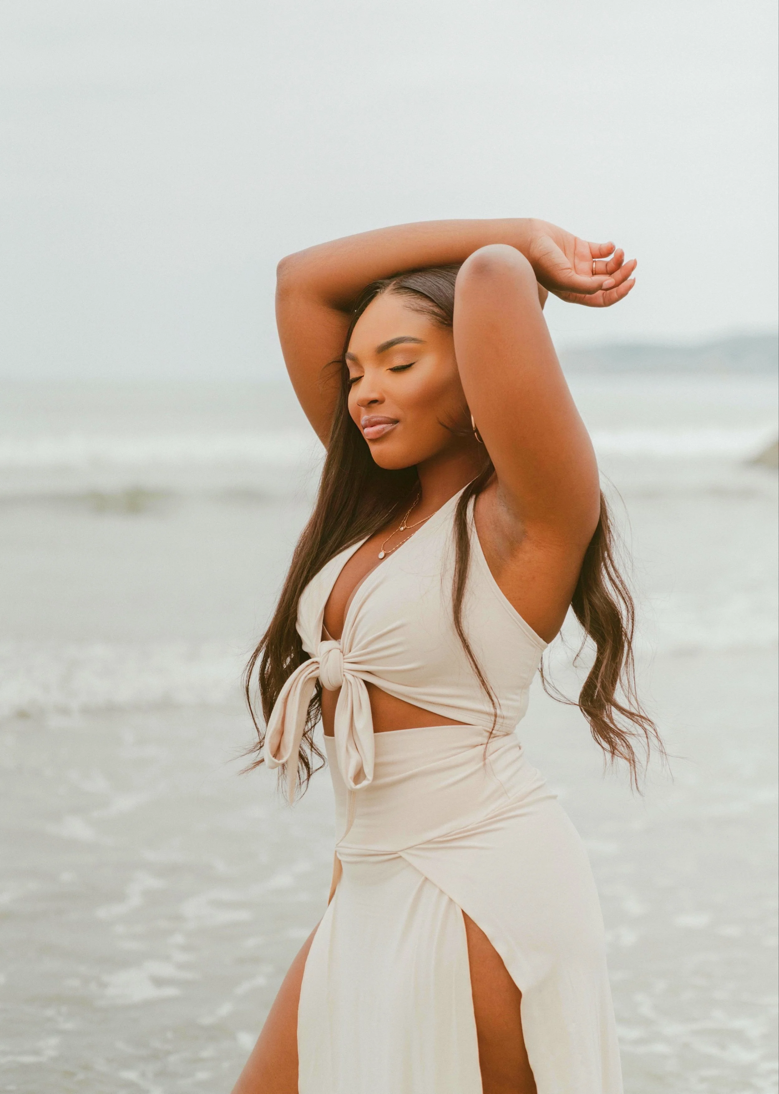 A woman in a beige dress with a front knot and side slit is standing on the beach with her eyes closed, with the ocean in the background.
