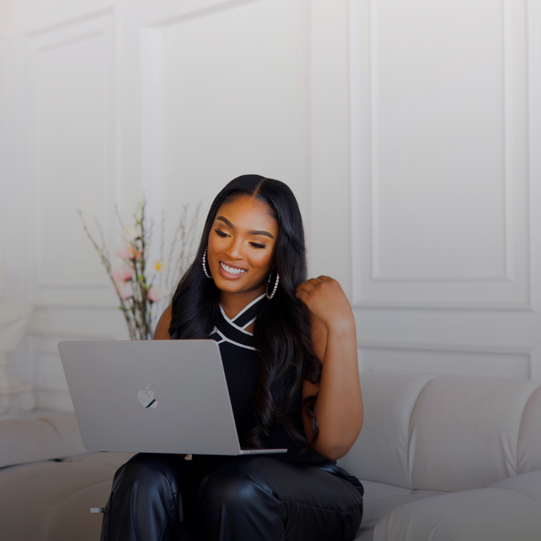 A woman sitting on a white couch, using a silver laptop, smiling, with a vase of pink and white flowers behind her in a white paneled room.