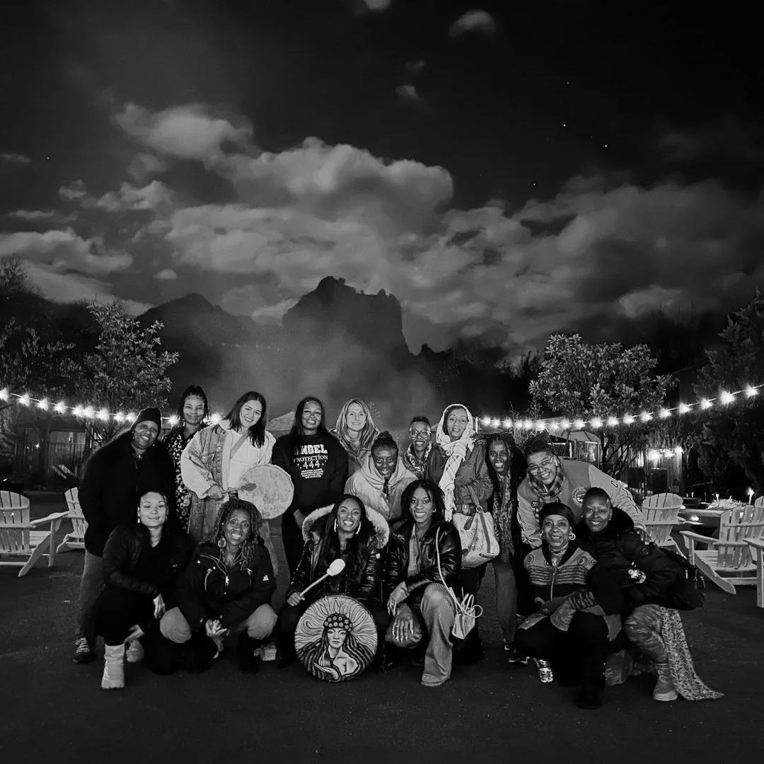 A group of 15 diverse people smiling at night outdoors, some kneeling and some standing, with string lights overhead and mountains in the background.