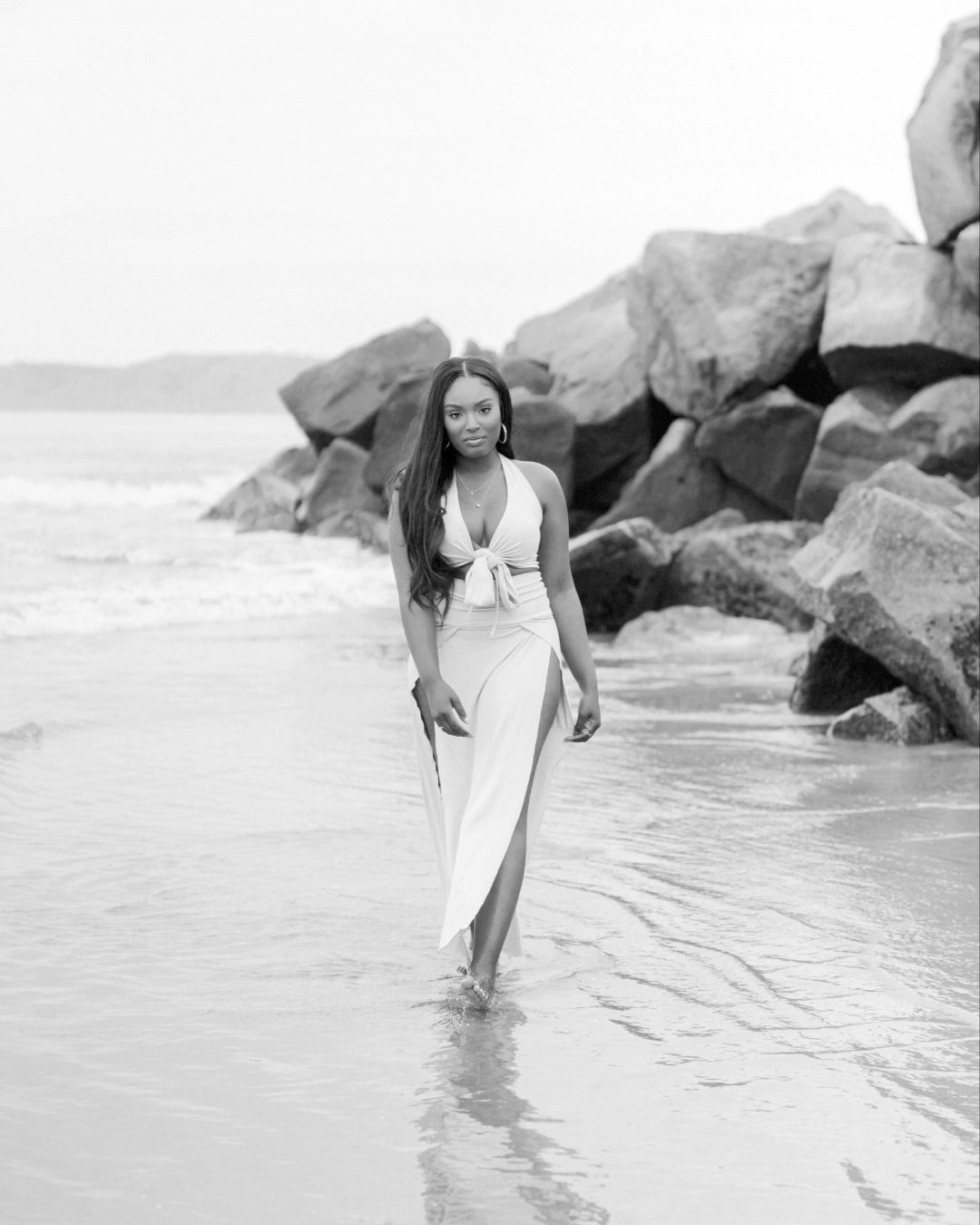 A woman walking on the beach wearing a long slit dress with rocks in the background.