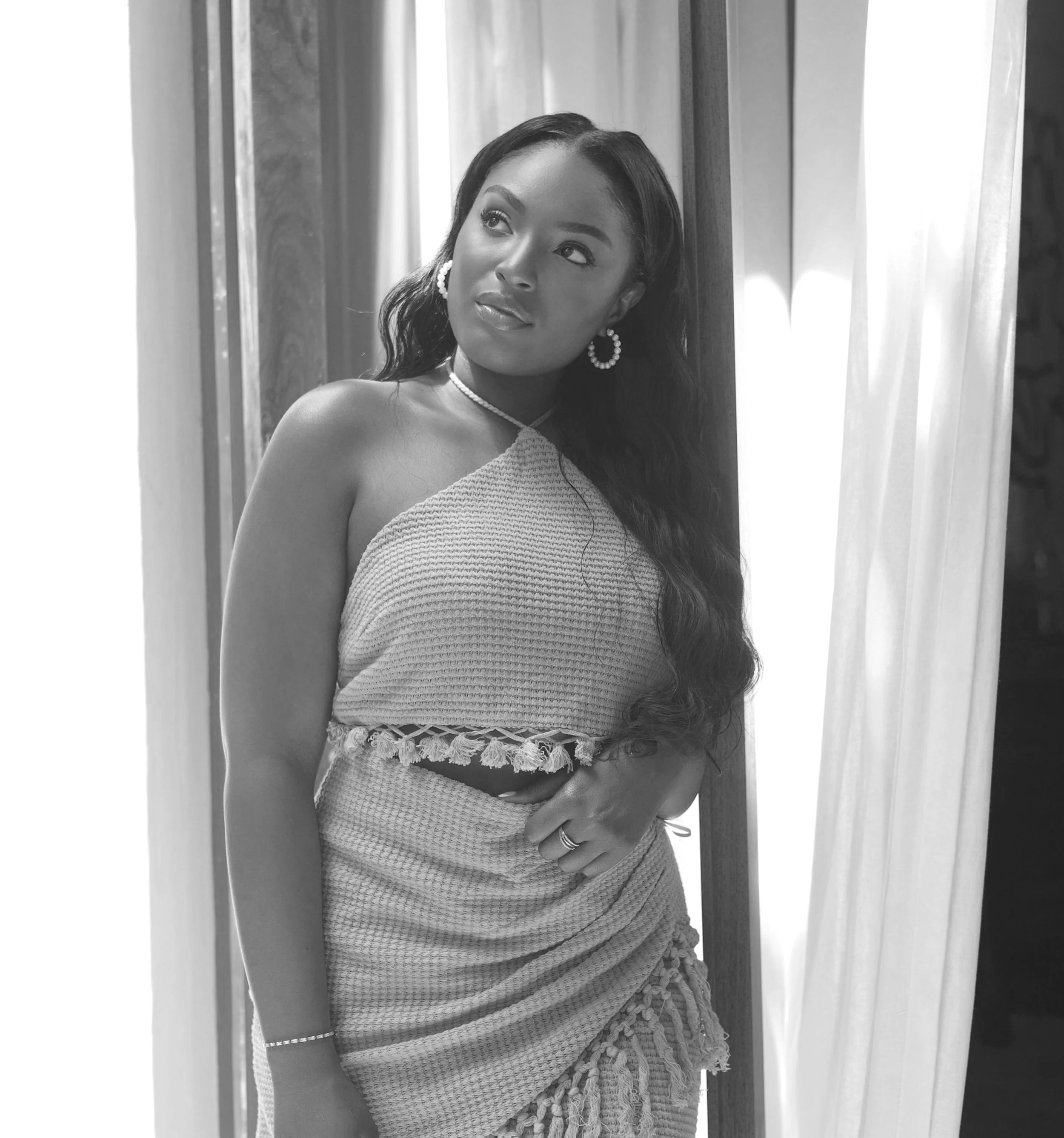 A young woman with long wavy hair standing near a curtain with soft lighting, wearing a sleeveless textured dress, earrings, a necklace, and a bracelet, looking away thoughtfully.