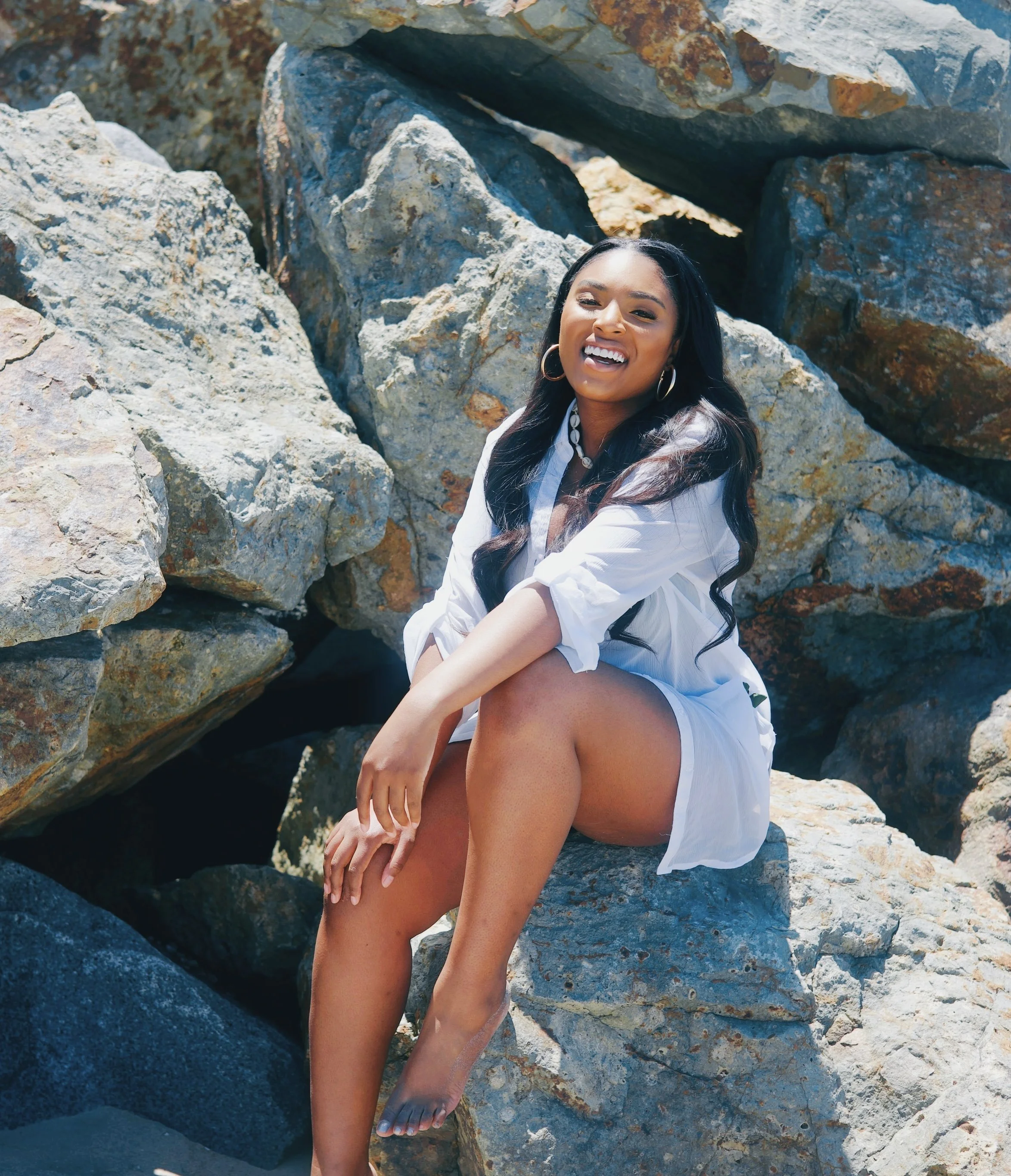 A woman sitting on rocks near the beach, smiling and wearing a white dress, with long dark hair and hoop earrings.