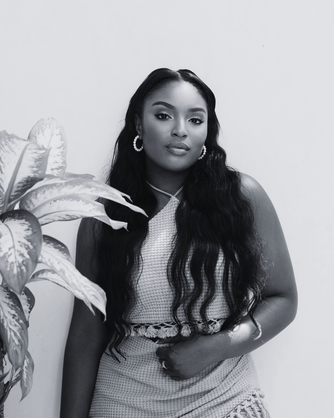 A woman with long wavy hair wearing hoop earrings and a sleeveless top standing next to a potted plant, posed in front of a plain wall.