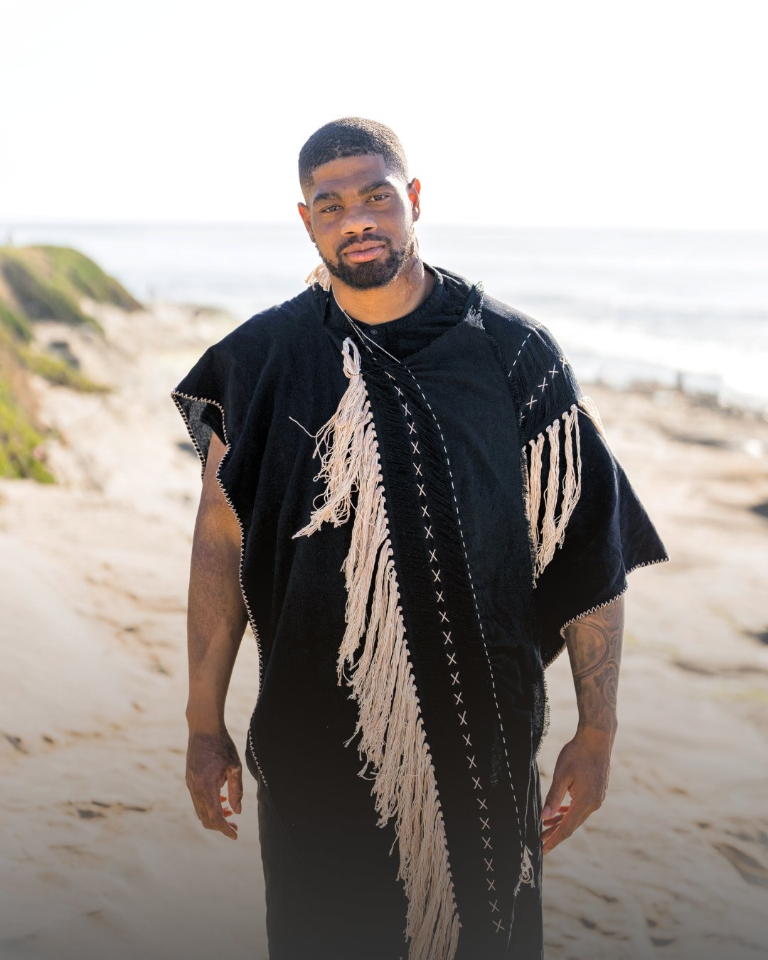 A man standing on a sandy beach near the ocean, wearing a black garment with cream-colored fringe and decorative stitching.