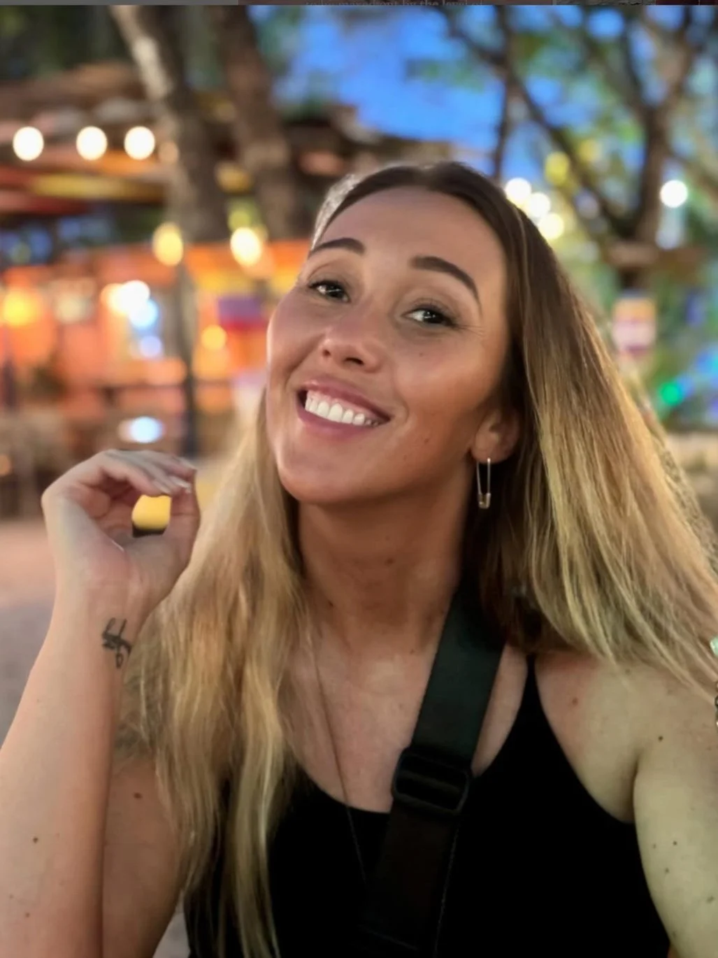 A woman with long blonde hair smiling at a lively outdoor setting at dusk with colorful lights and trees in the background.