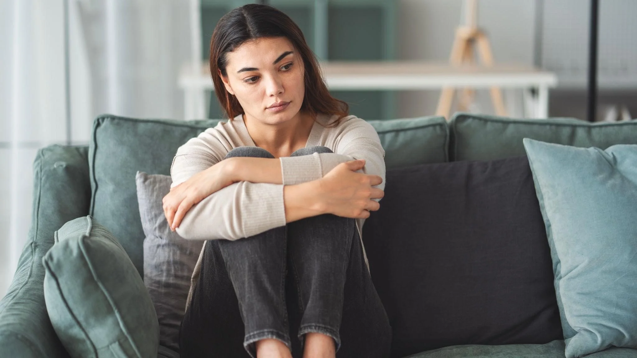 A woman sitting on a couch hugging her knees, with a sad or contemplative expression.