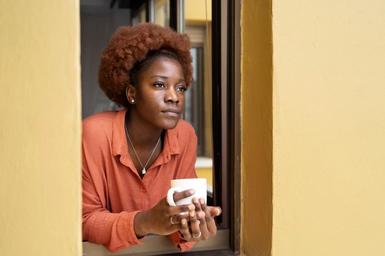 A woman with curly red hair sitting at a window sill, holding a white mug, looking thoughtfully outside, wearing an orange shirt.