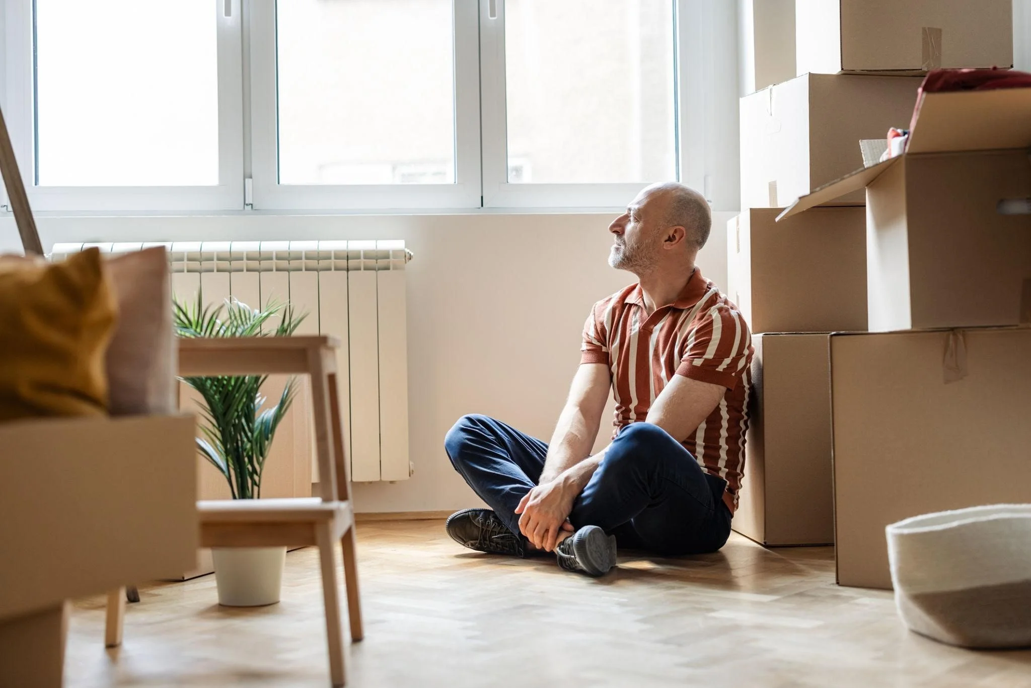 A man sitting on the floor amidst cardboard boxes in a bright room with large windows, plants, and wooden flooring.