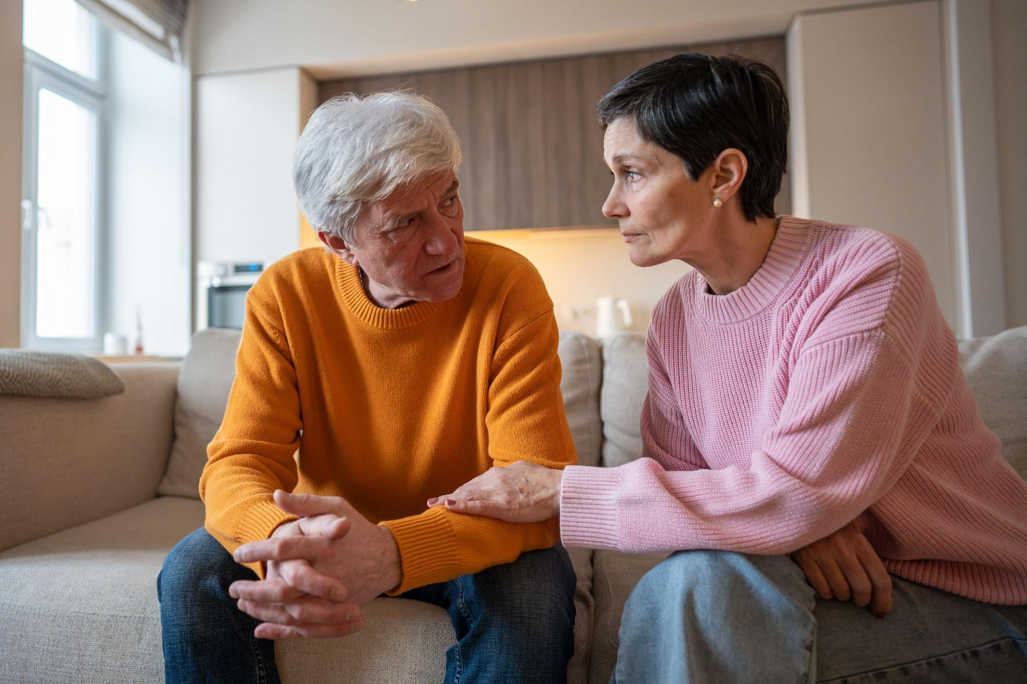 An older man and woman are sitting close together on a sofa, engaged in a serious conversation. They are holding hands and looking into each other's eyes in a cozy, well-lit living room.