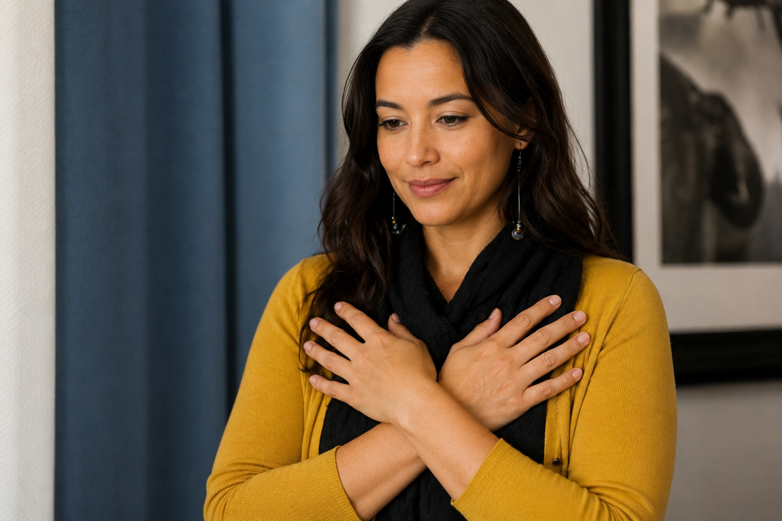 A woman in a mustard-colored sweater and black scarf standing indoors, smiling softly with her eyes closed and hands crossed over her chest.