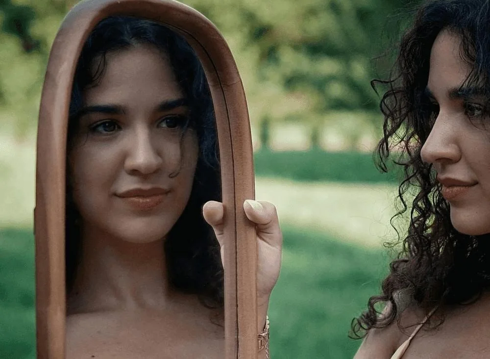 A woman with dark, curly hair looks at her reflection in a handheld mirror outdoors in a green park.