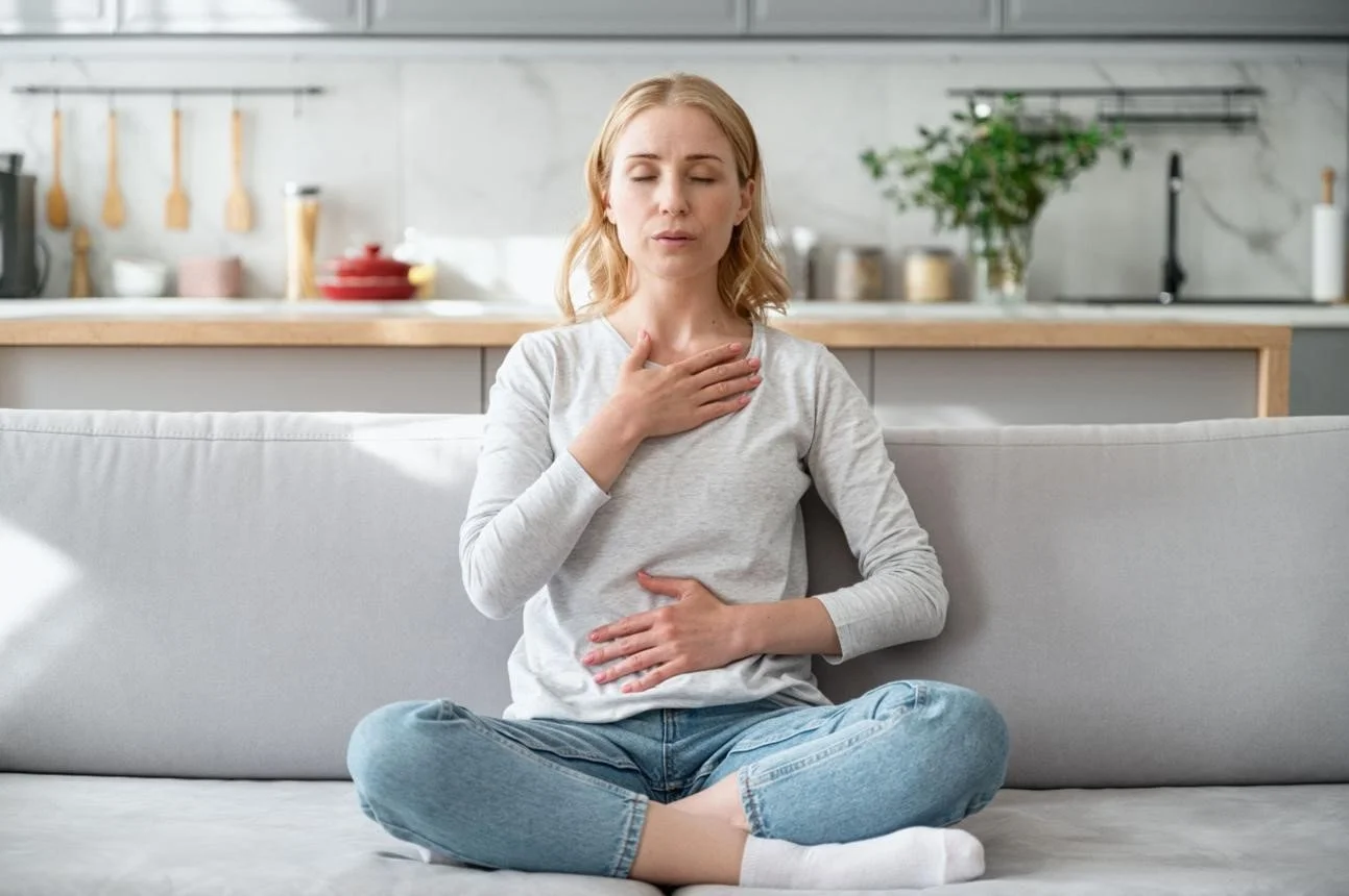 A woman sitting cross-legged on a couch with her eyes closed, holding her throat and stomach, appearing to be experiencing discomfort or distress.