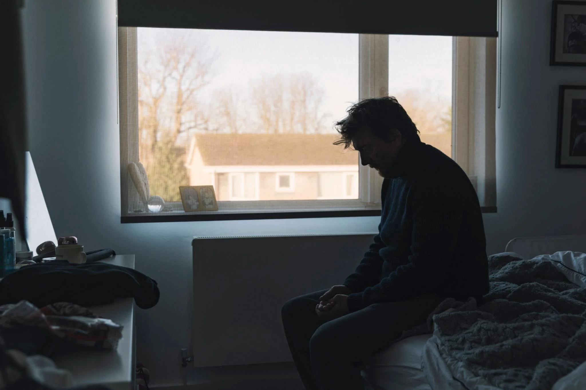 A person with dark hair and a beard sitting on a bed, facing downward, in a dimly lit room with sunlight coming through a window. The room has photos and various items on the walls and a dresser.