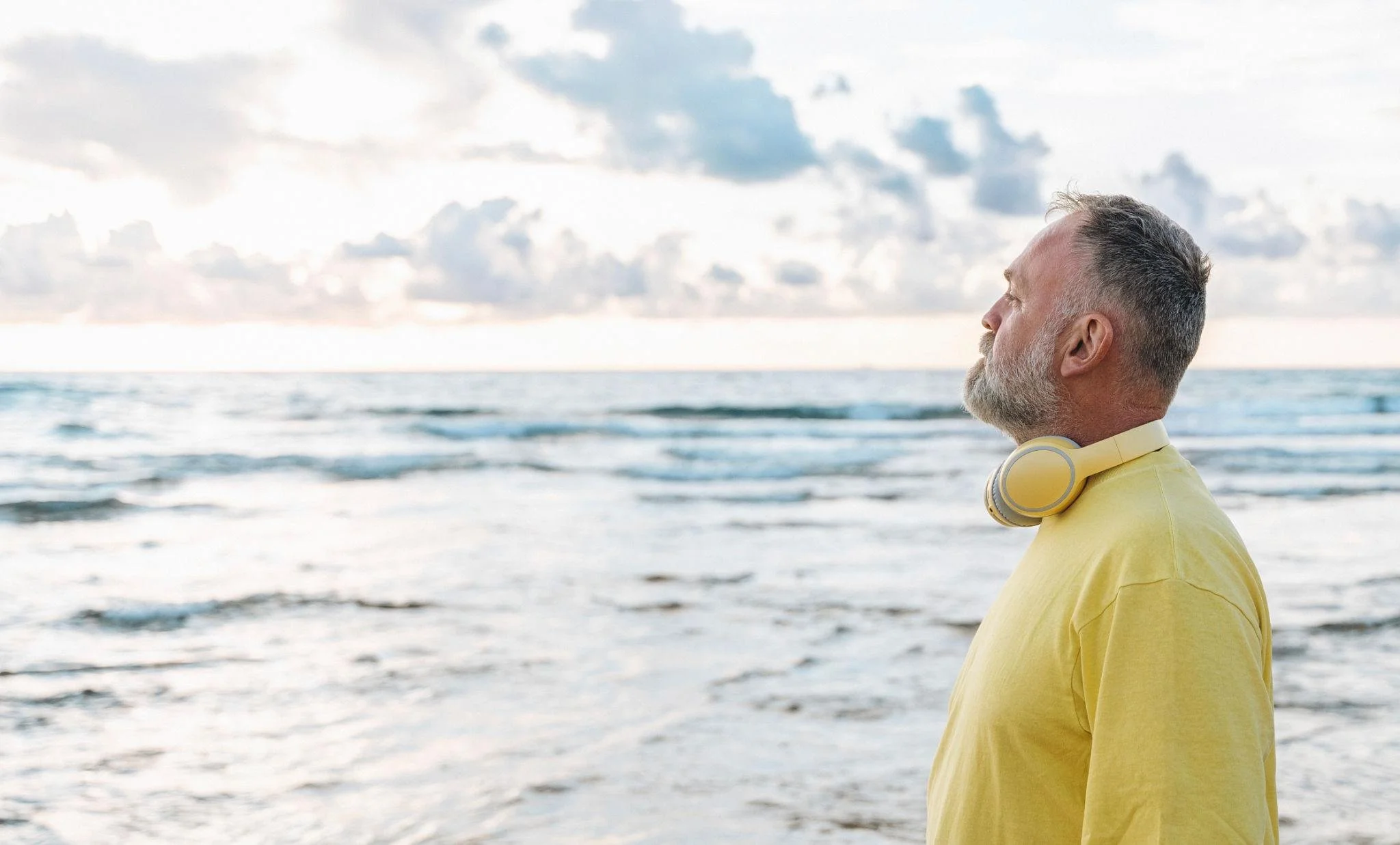 A man with gray hair and beard wearing a yellow shirt and headphones around his neck sitting on the beach, looking at the ocean and sky.