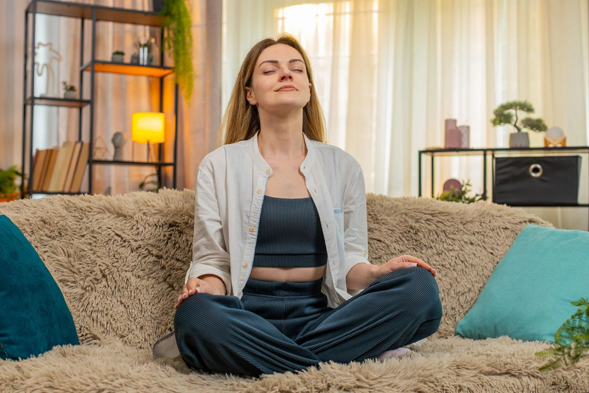 A woman with long brown hair, wearing a white shirt and black sports bra, is sitting cross-legged on a plush beige rug in a bright room, meditating with eyes closed and a peaceful expression.