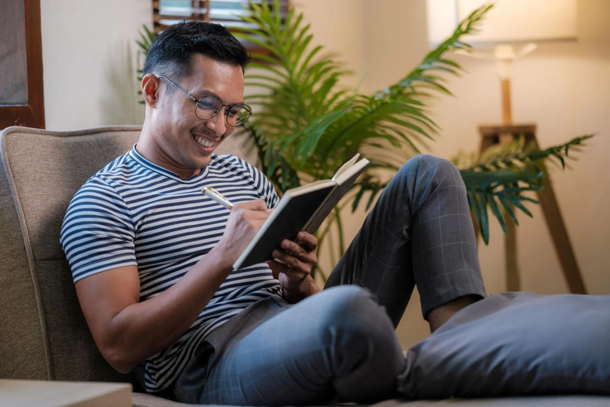 A man sitting on a beige couch, smiling while reading a book, with a large green plant and a small table with a lamp in the background.