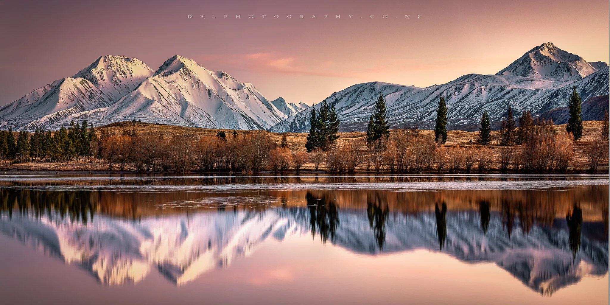 Snow-capped mountains reflecting in a calm lake with trees and shrubs along the shoreline at sunset.