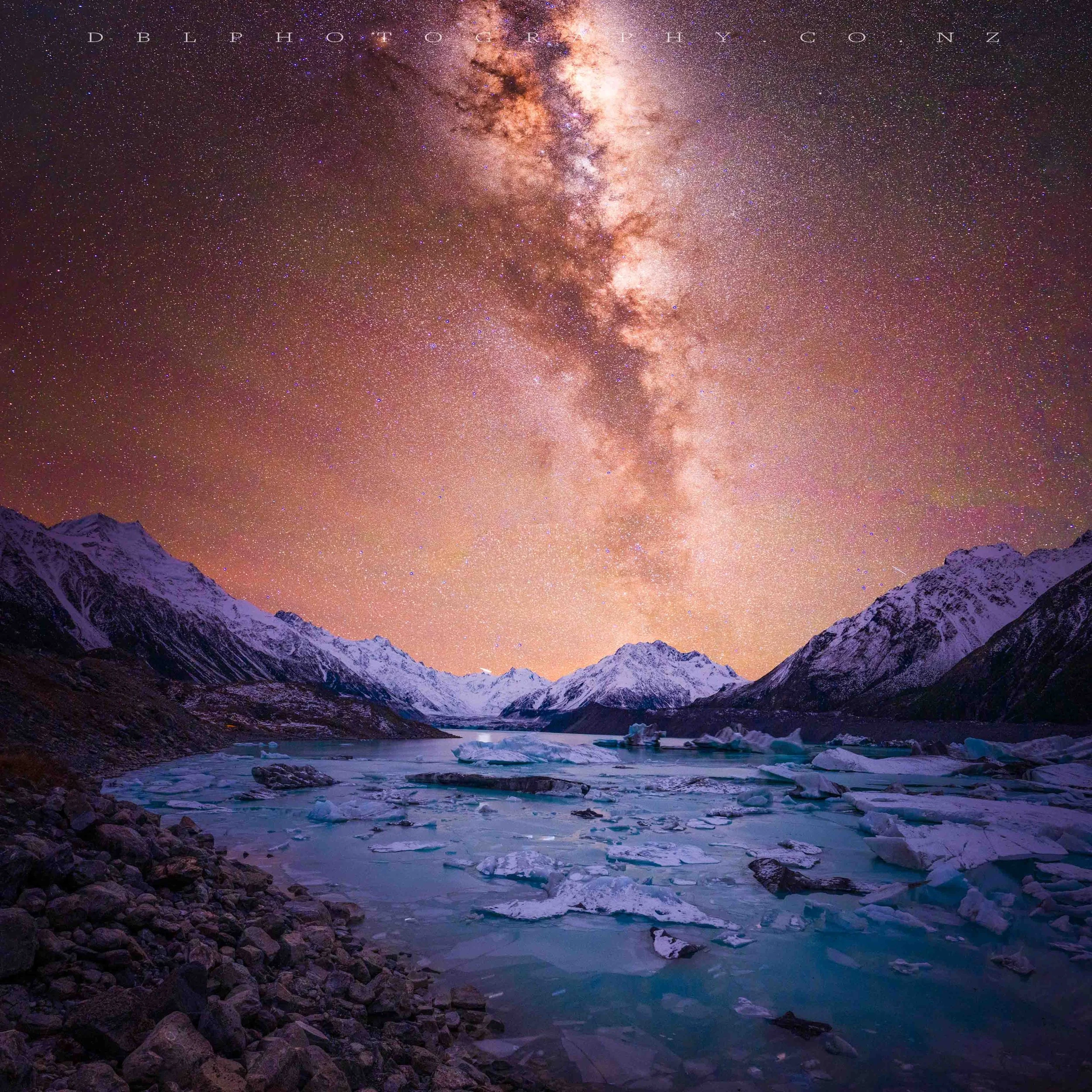 Nighttime landscape of snow-covered mountains, ice floating on a partially frozen river, and a star-filled sky with the Milky Way galaxy visible overhead.