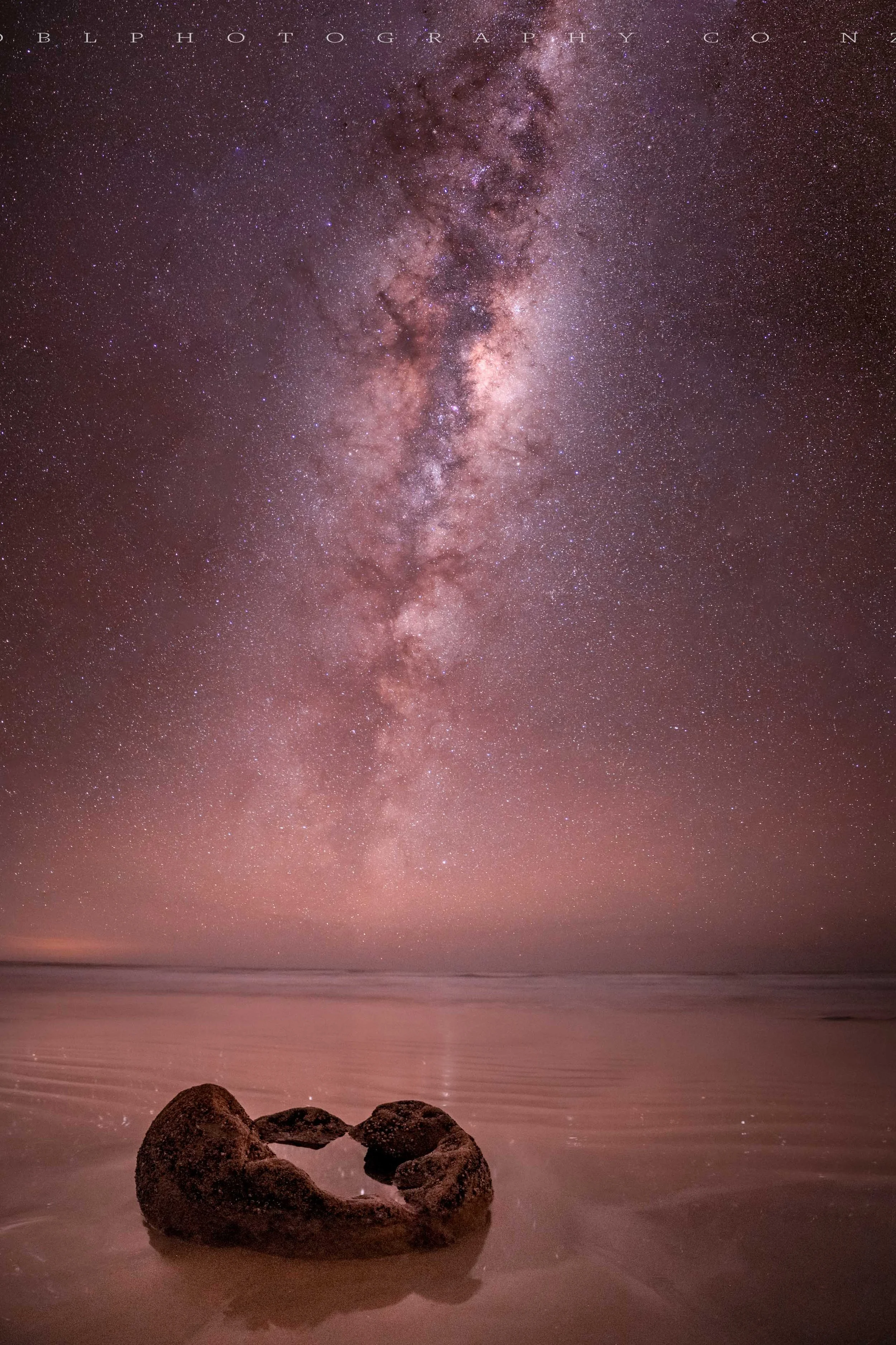 A star-filled night sky with the Milky Way galaxy visible, reflected in calm ocean waters with a heart-shaped rock formation in the foreground.