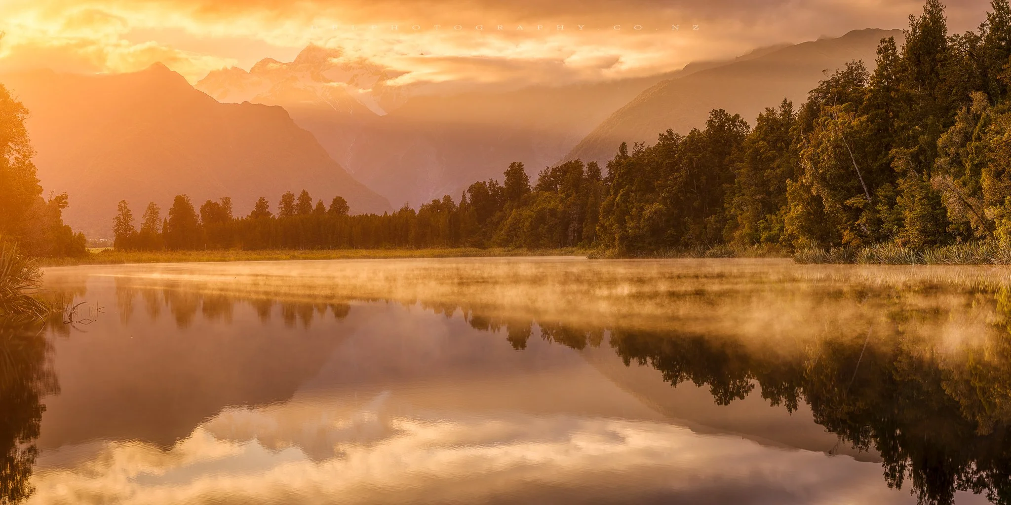 Sunset over mountains and a river with mist rising, surrounded by trees and dense forest.