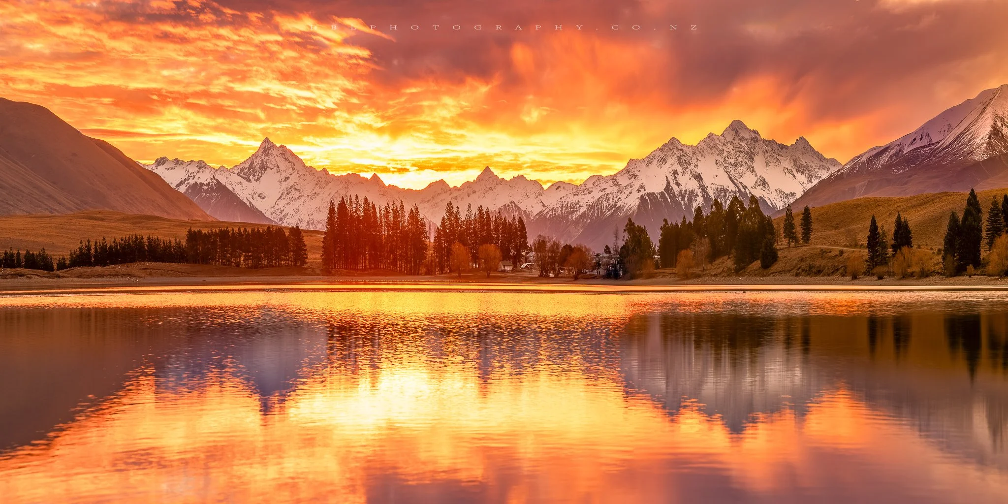 Sunset over snow-capped mountains reflecting on a calm lake with trees along the shoreline.