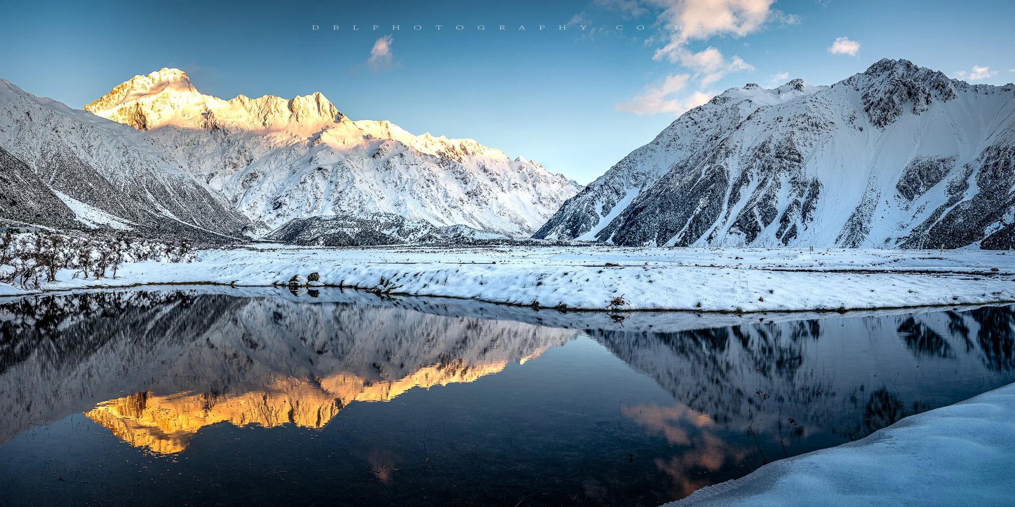 Snow-covered mountains reflect in a calm river, with a partly cloudy sky above.