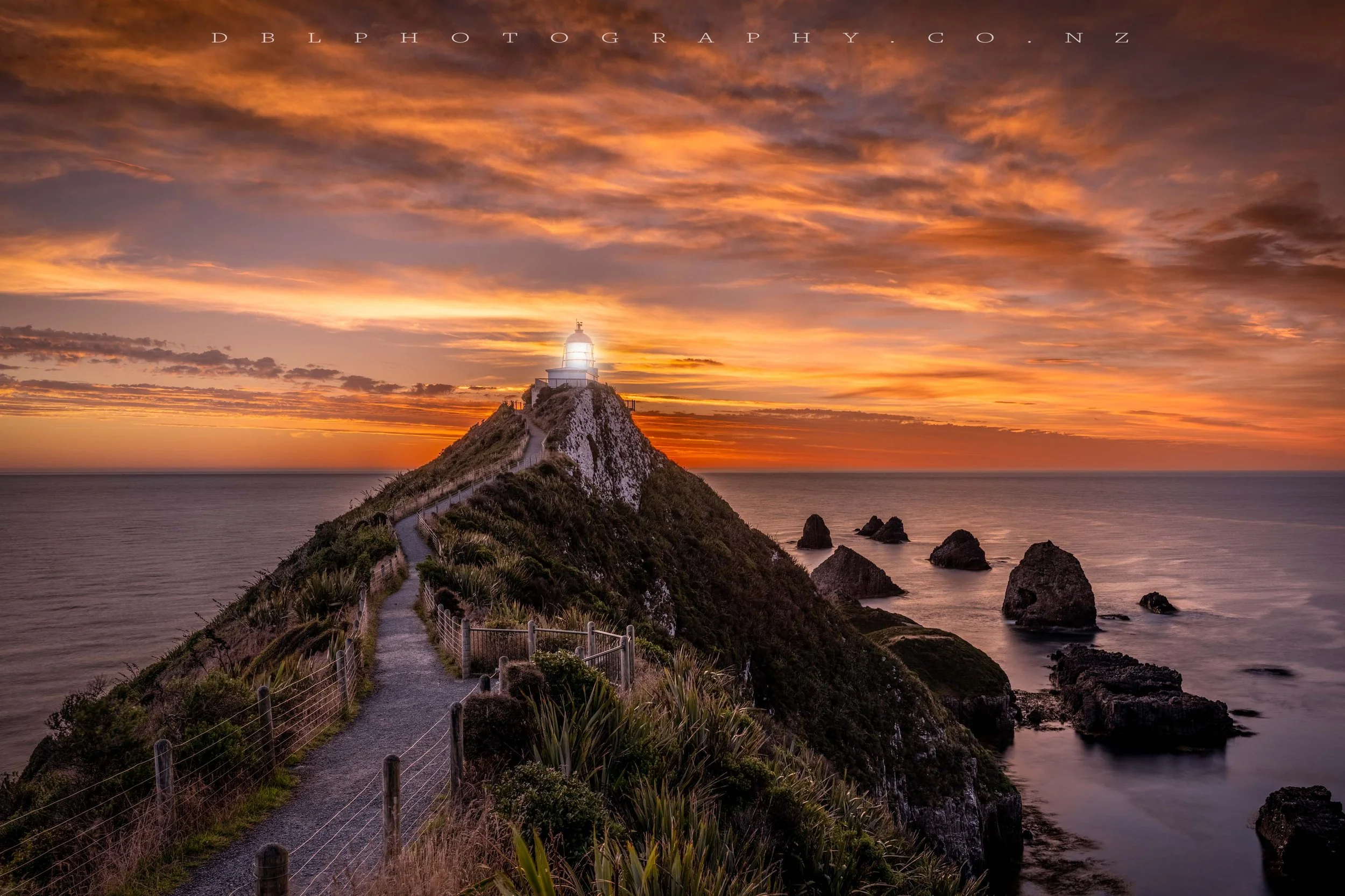A scenic view of a lighthouse on a cliff at sunset, with a winding path leading to the lighthouse and rocks in the ocean below.