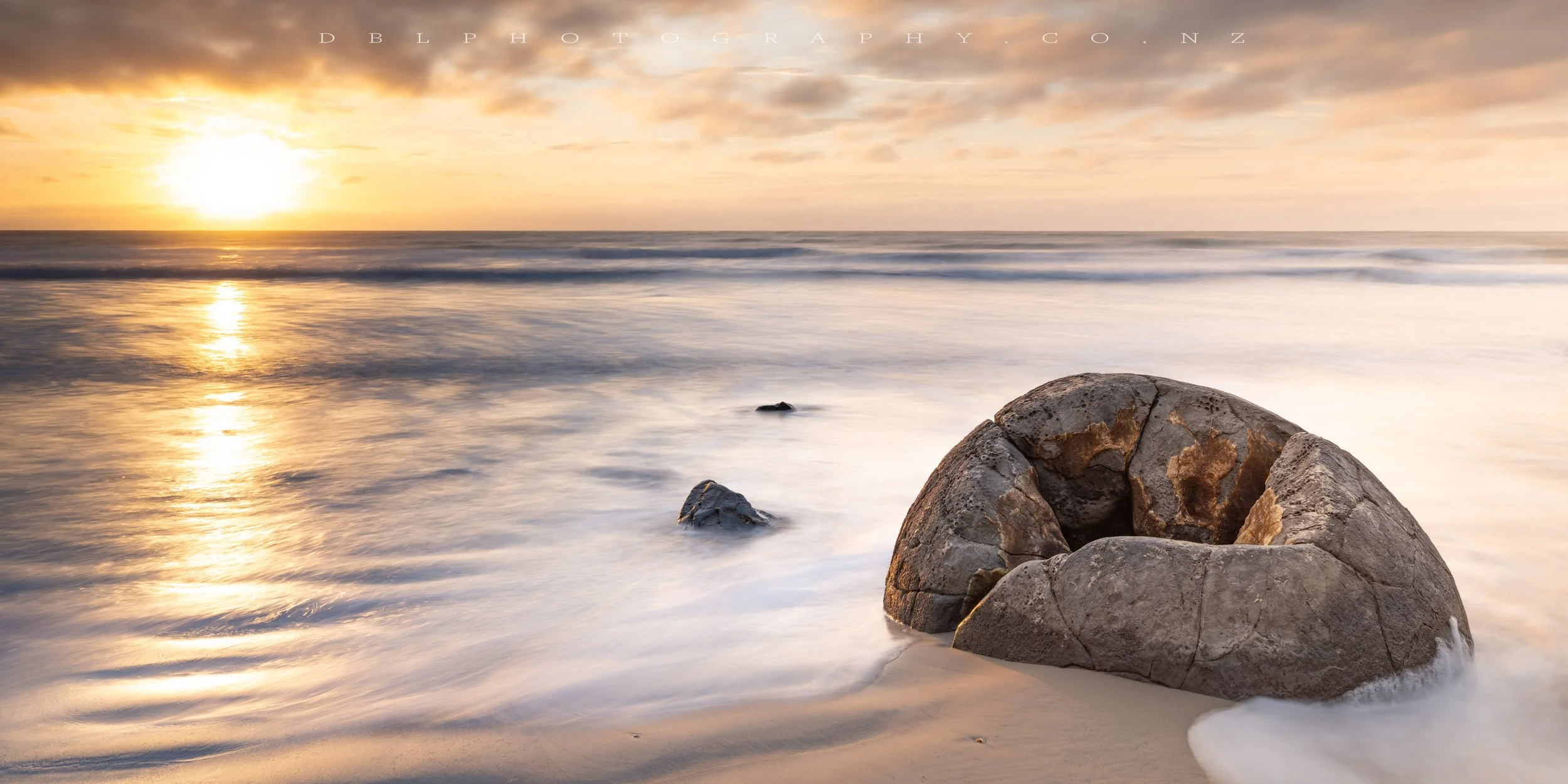 A large rounded rock partially submerged on a sandy beach, with smaller rocks nearby, during a sunset with orange and yellow hues, and gentle waves reflecting the sky.