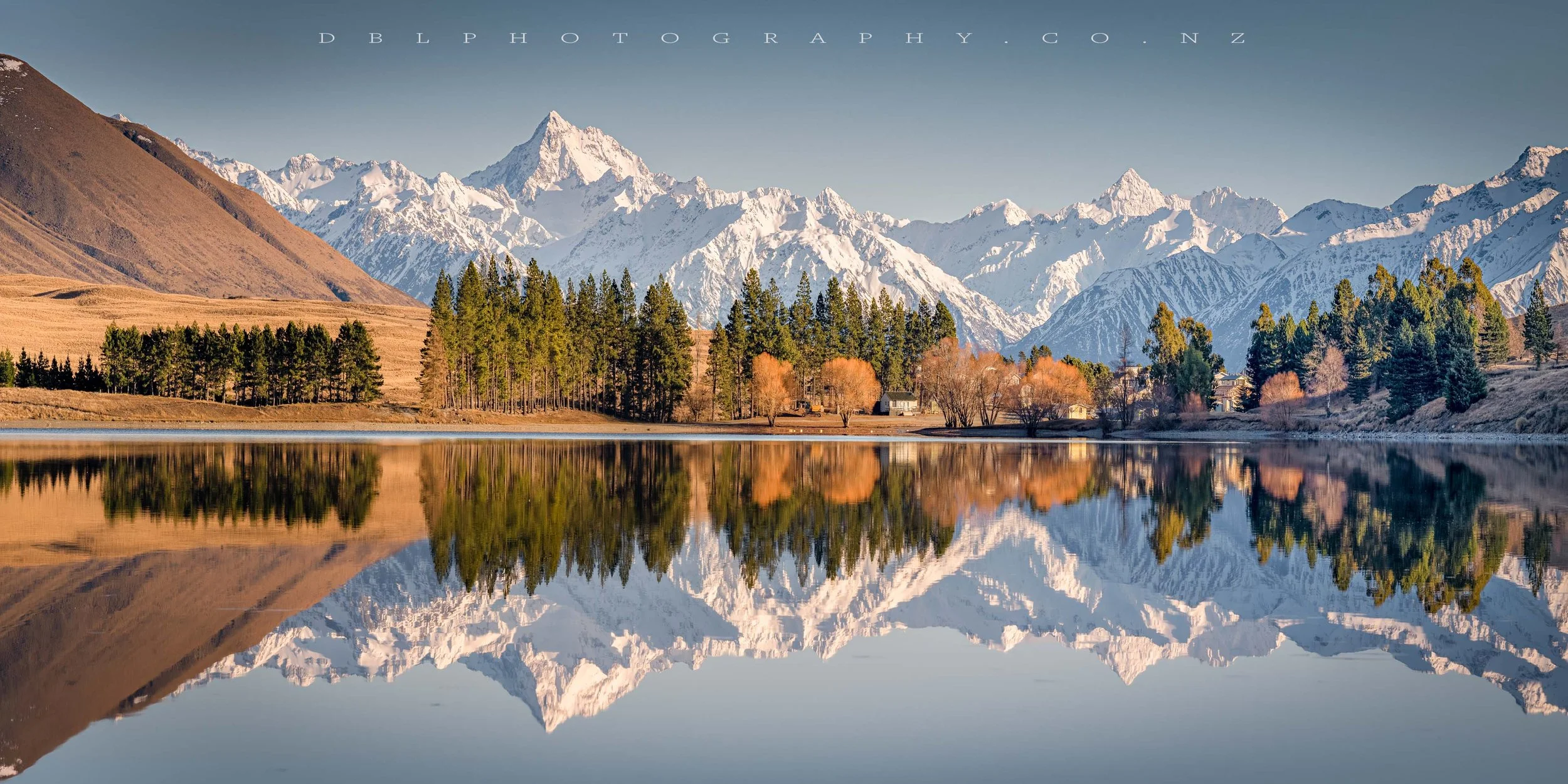 Snow-capped mountains over a lake with trees and a few houses, reflecting in the water.