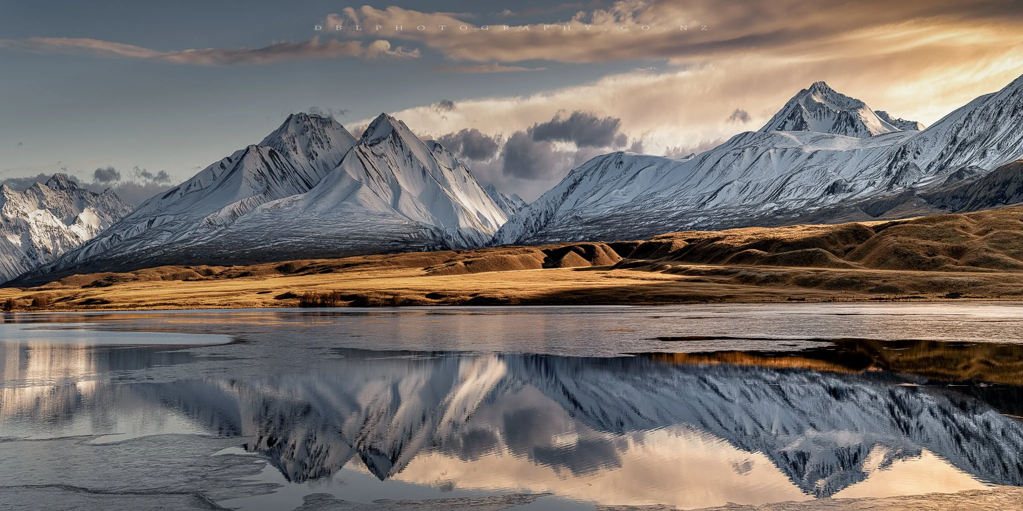 Snow-covered mountains reflecting in a lake with grassy hills in the foreground and a partly cloudy sky.