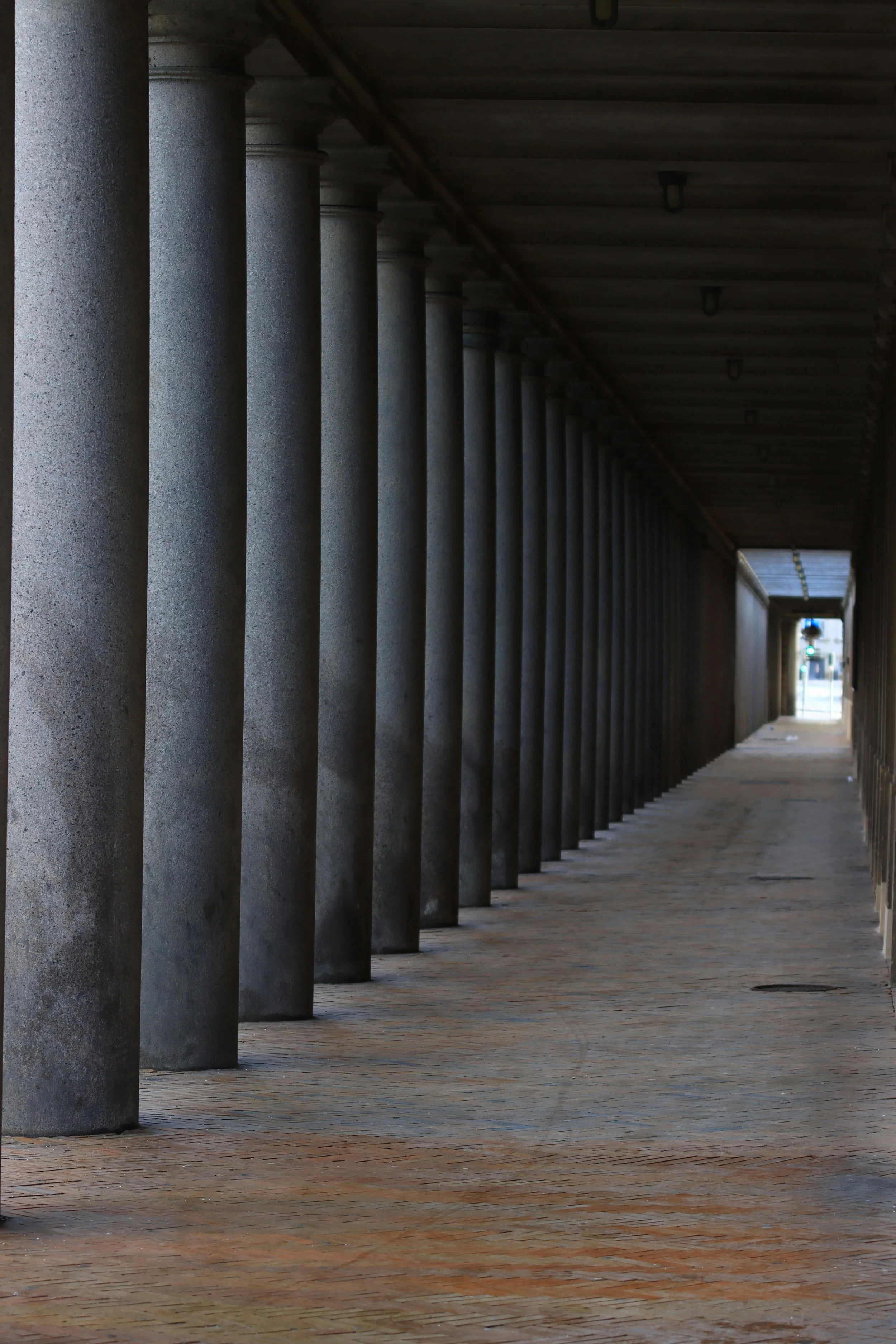 A covered walkway with evenly spaced concrete columns on the left side and a brick-paved floor. The walkway extends into the distance, ending in a brighter area with a green traffic light and street scene visible at the far end.
