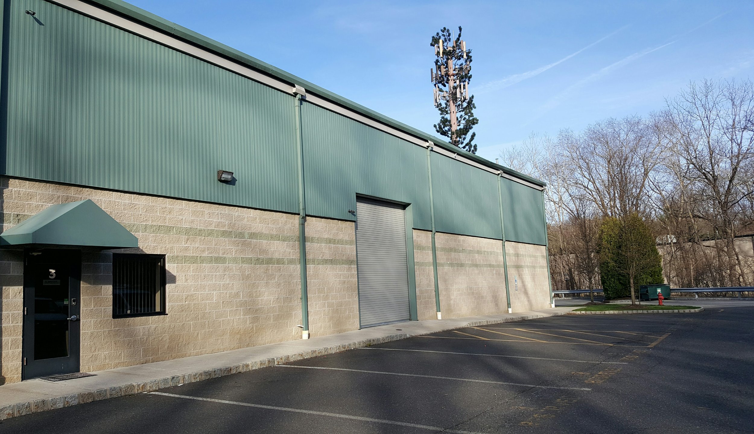 Exterior view of a commercial building with a green metal upper section and beige brick lower section, a parking lot in front, and trees in the background.