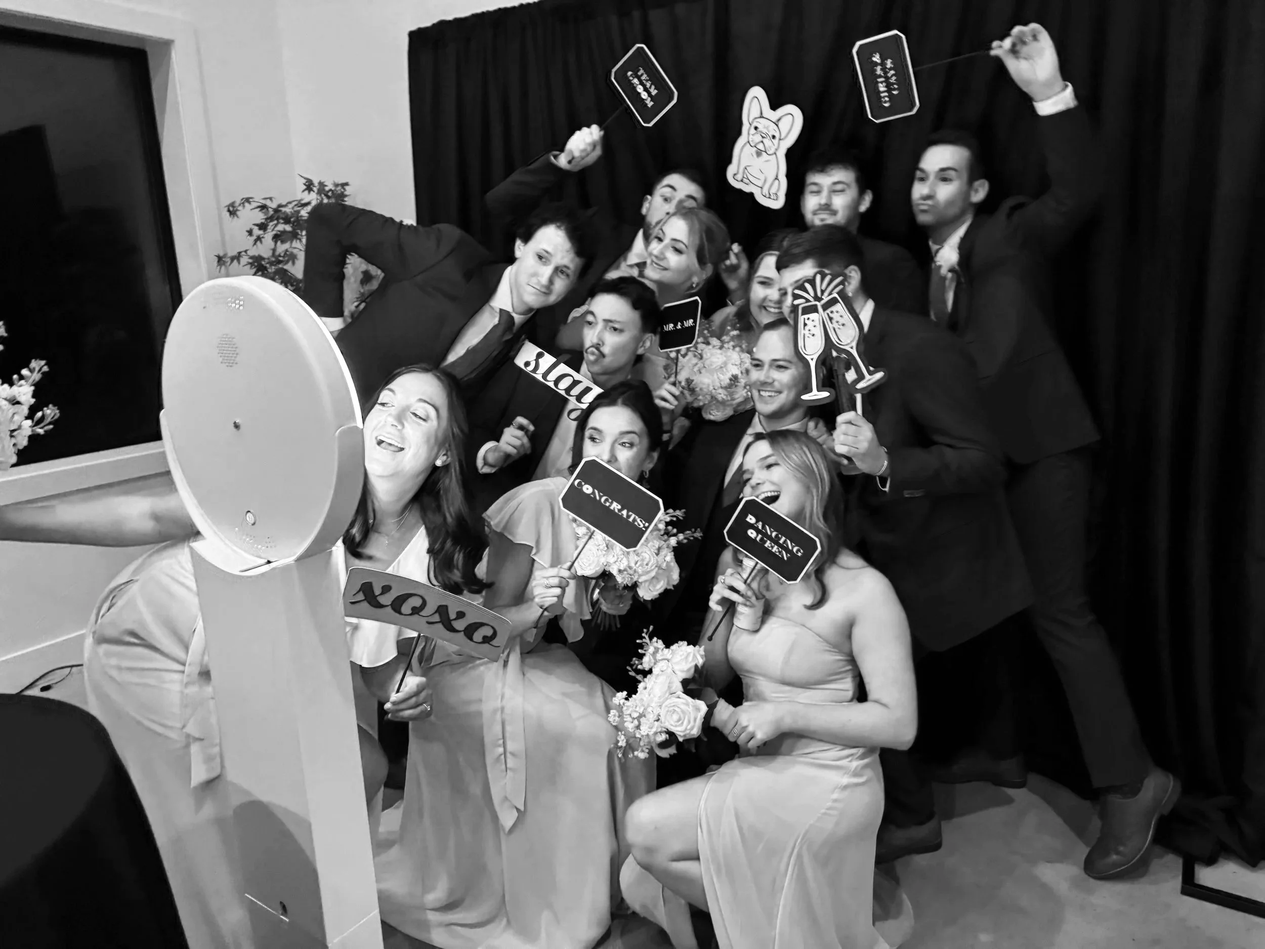 A group of people celebrating at a wedding or party, taking a selfie with photo props and signs like "congrats," "say yes," "dancing queen," and others, in front of a dark curtain backdrop.
