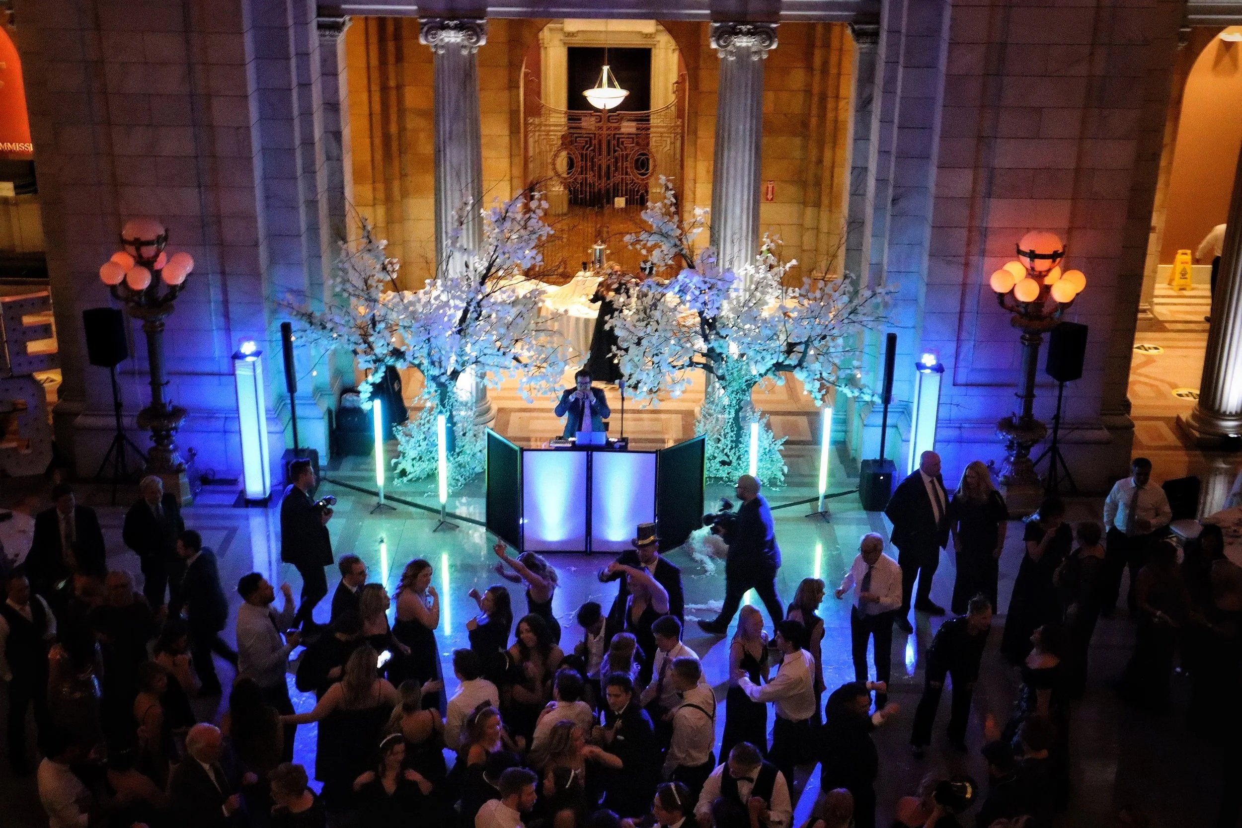 People dancing and socializing in a grand hall with a DJ booth at the center, decorated with illuminated artificial cherry blossom trees and colorful lighting, in an elegant building with large columns and ornate architecture.